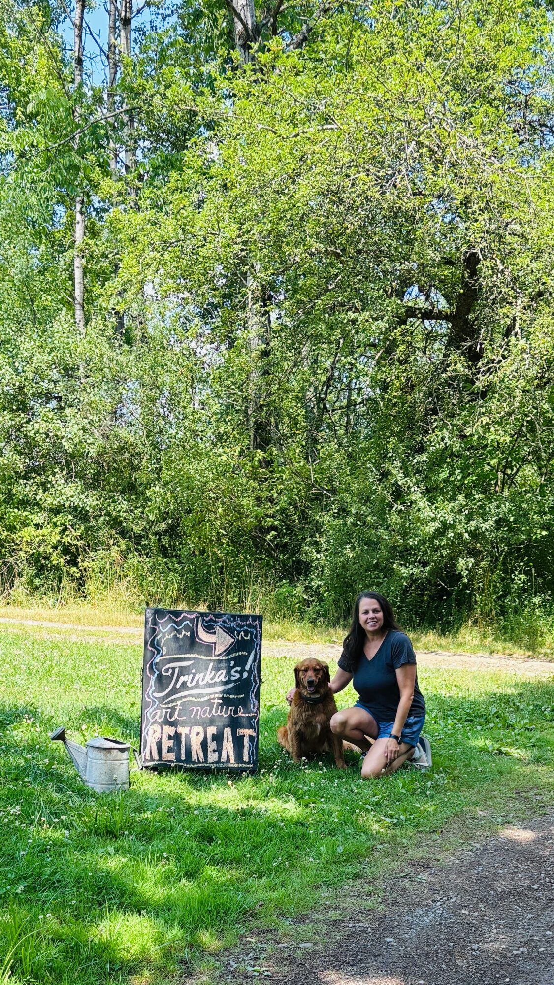 Person sitting on grass with a dog outdoors, trees in background, sunny day, signboard nearby.