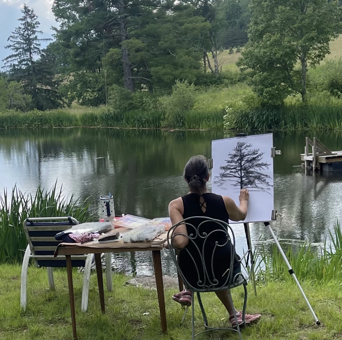 Person painting a tree on a canvas by a lake with trees and grass in the background.