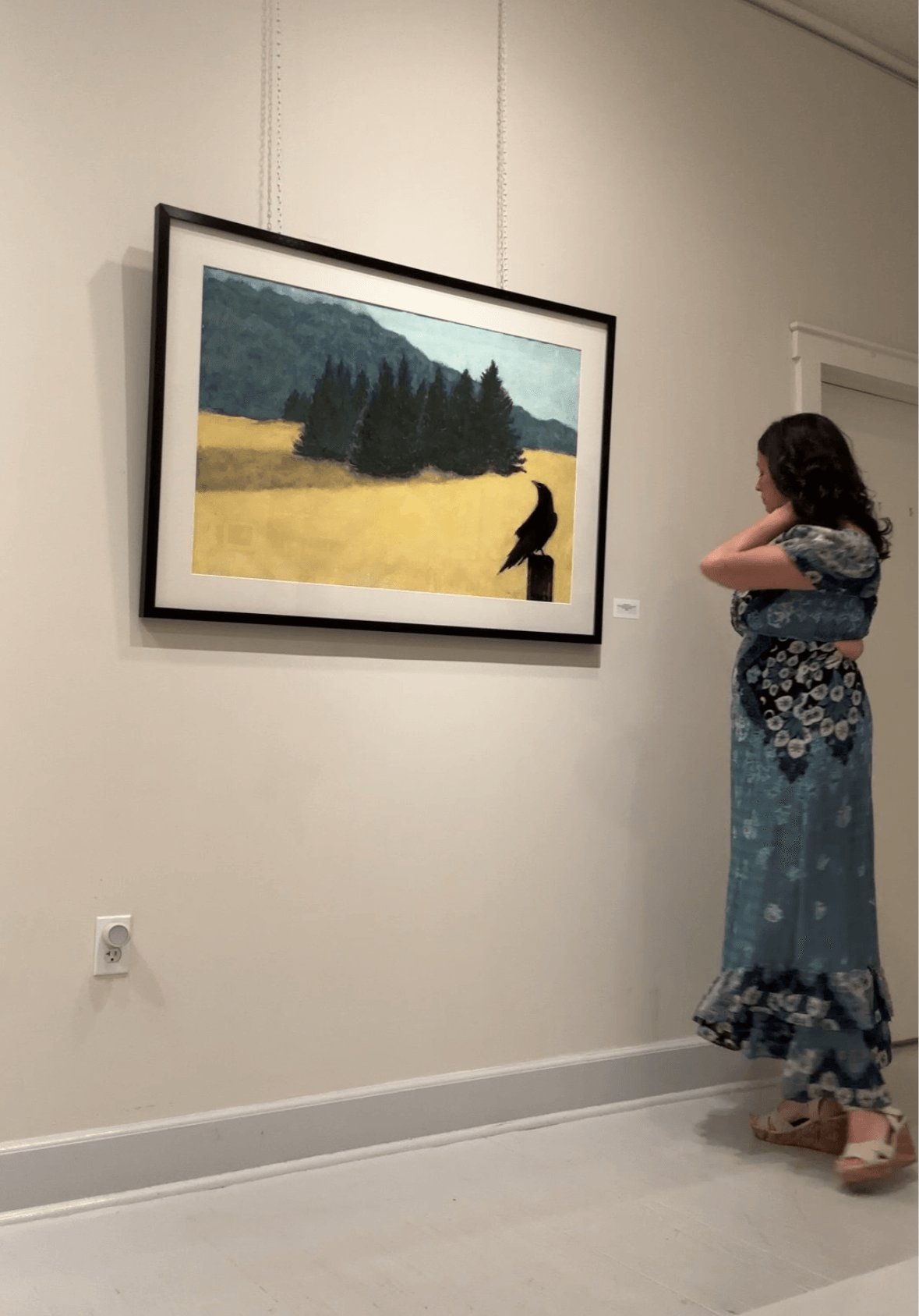 Woman in a floral dress looking at a landscape painting in a gallery.