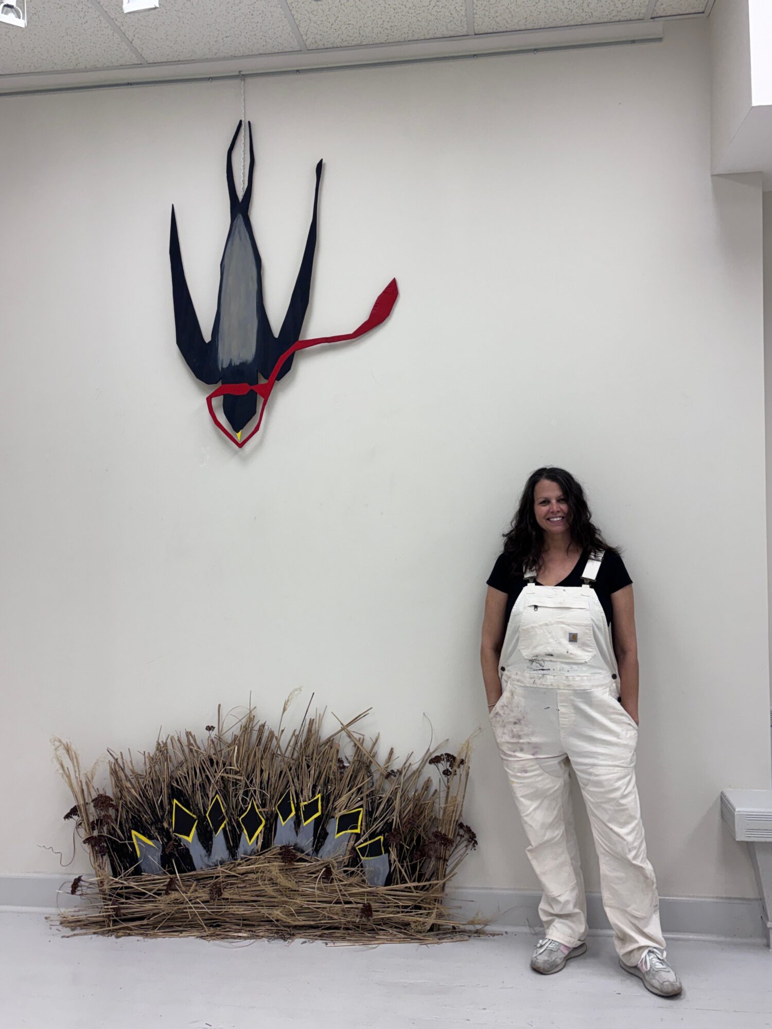 Person standing next to a wall-mounted sculpture of a rabbit with a red ribbon, and a pile of dried plants with black and yellow objects.