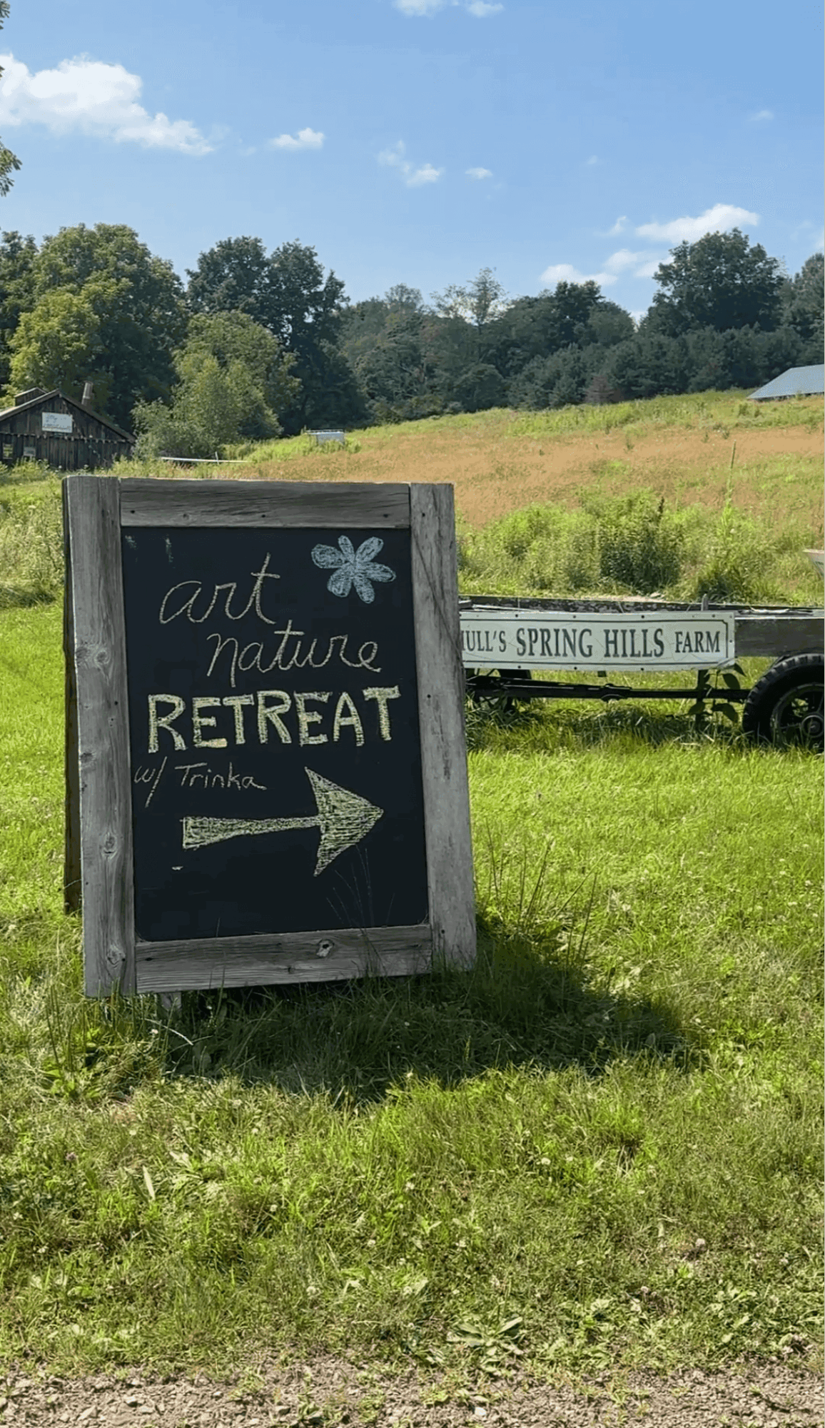 Chalkboard sign with arrow pointing right, advertising art, nature, and retreat, set on grassy field with trees and blue sky.