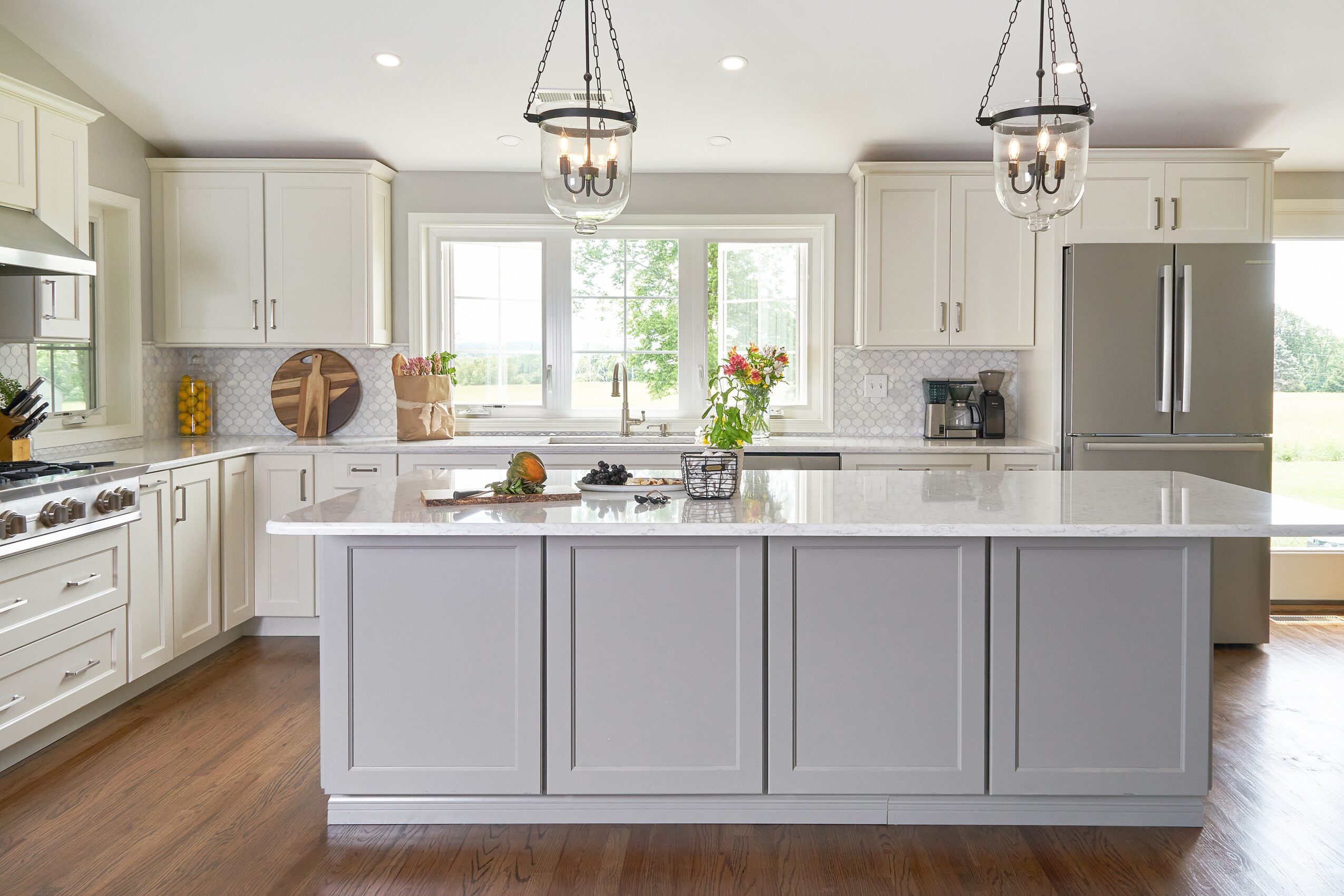 Bright kitchen with white cabinets, large island, window, and two pendant lights, with wooden floor and decorative items.