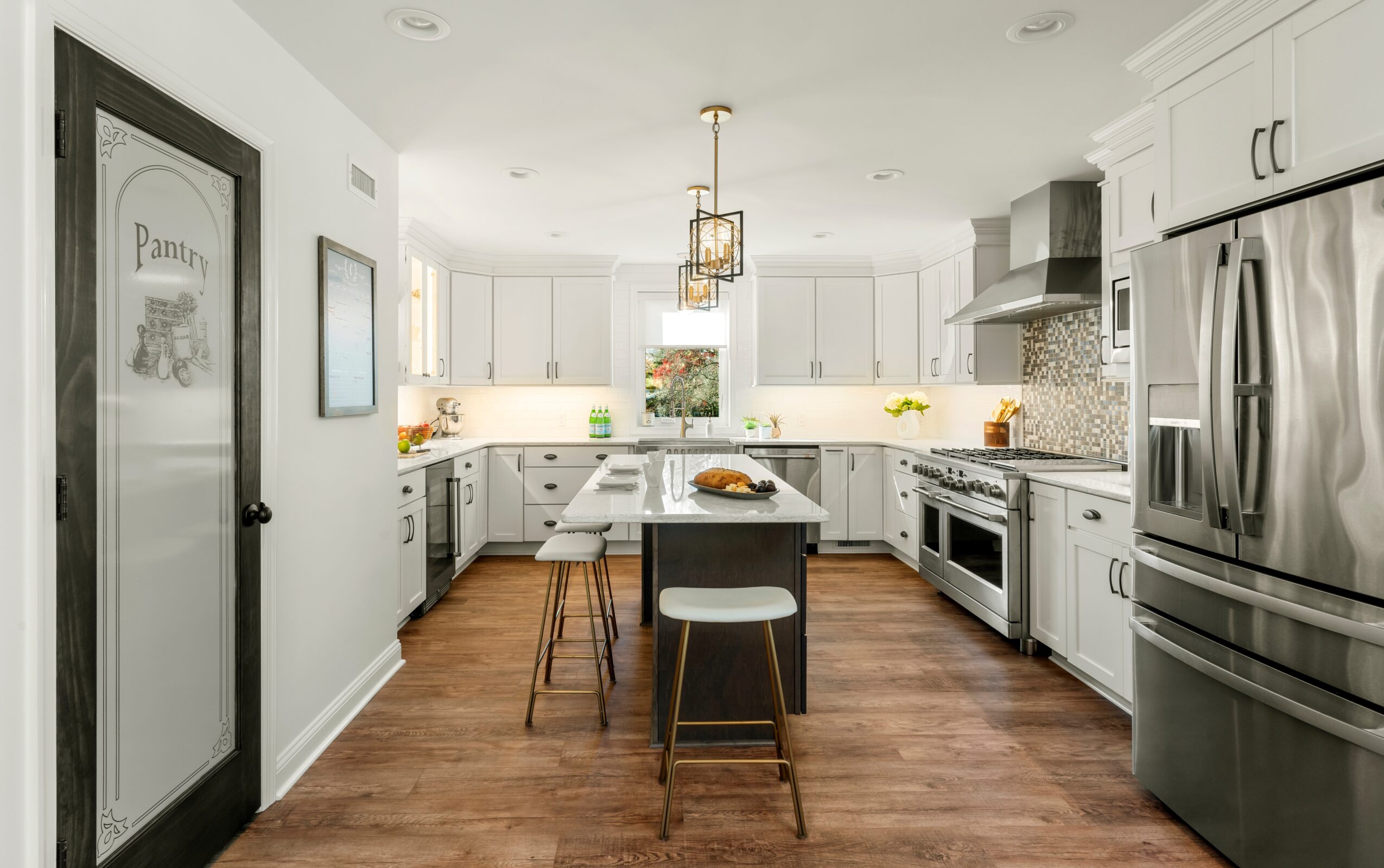 Modern kitchen with white cabinets, stainless steel appliances, and a central island with two stools, hardwood floors, and pendant lighting.