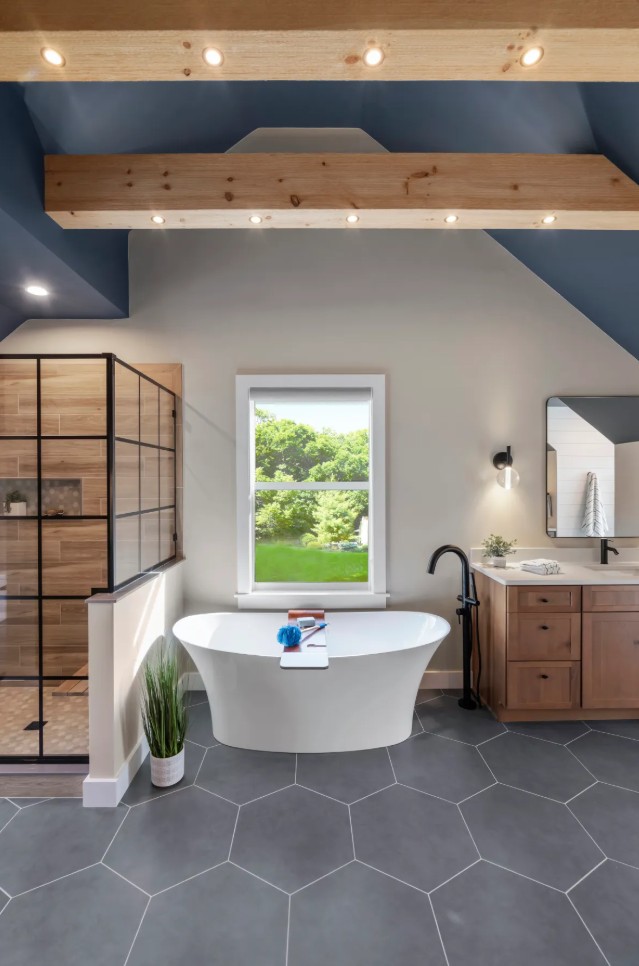 Bathroom with bathtub, window showing greenery, wooden cabinet, and gray hexagonal floor tiles.