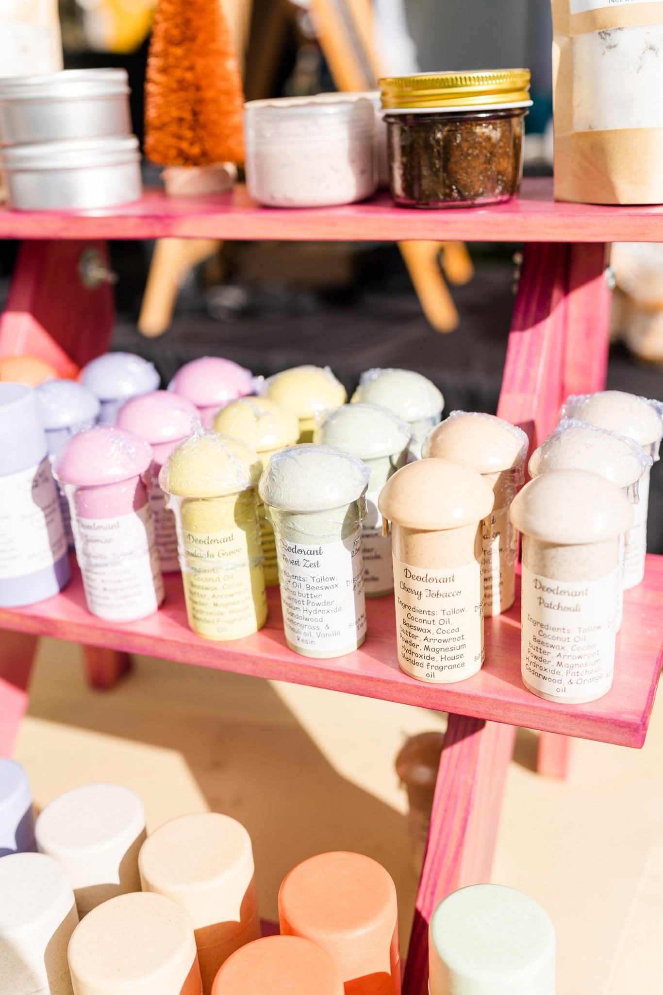 Colorful bath bombs and jars displayed on pink shelves at an outdoor market, with a blurred background.