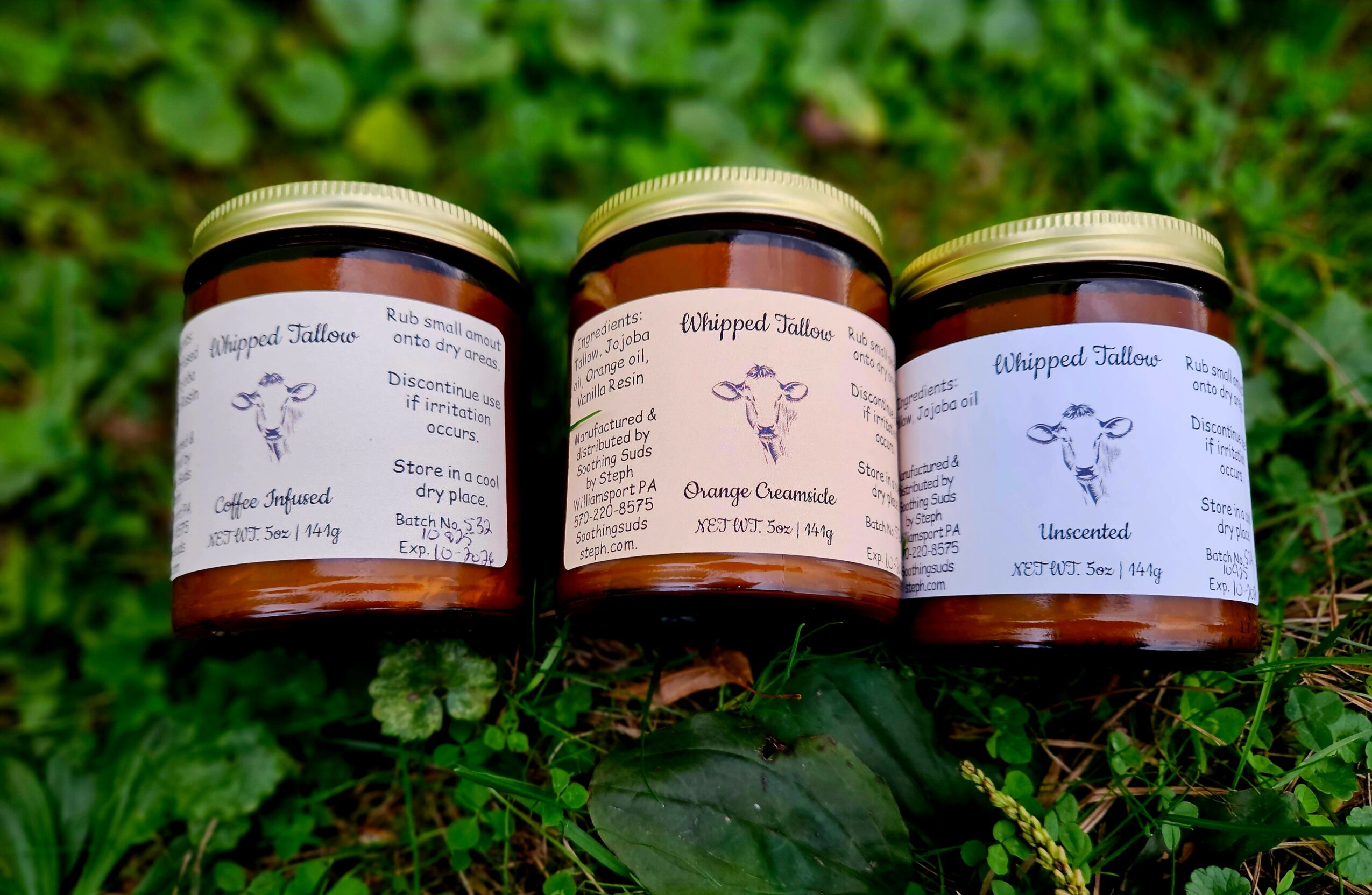 Three jars with labels are placed on grass with green foliage in the background.