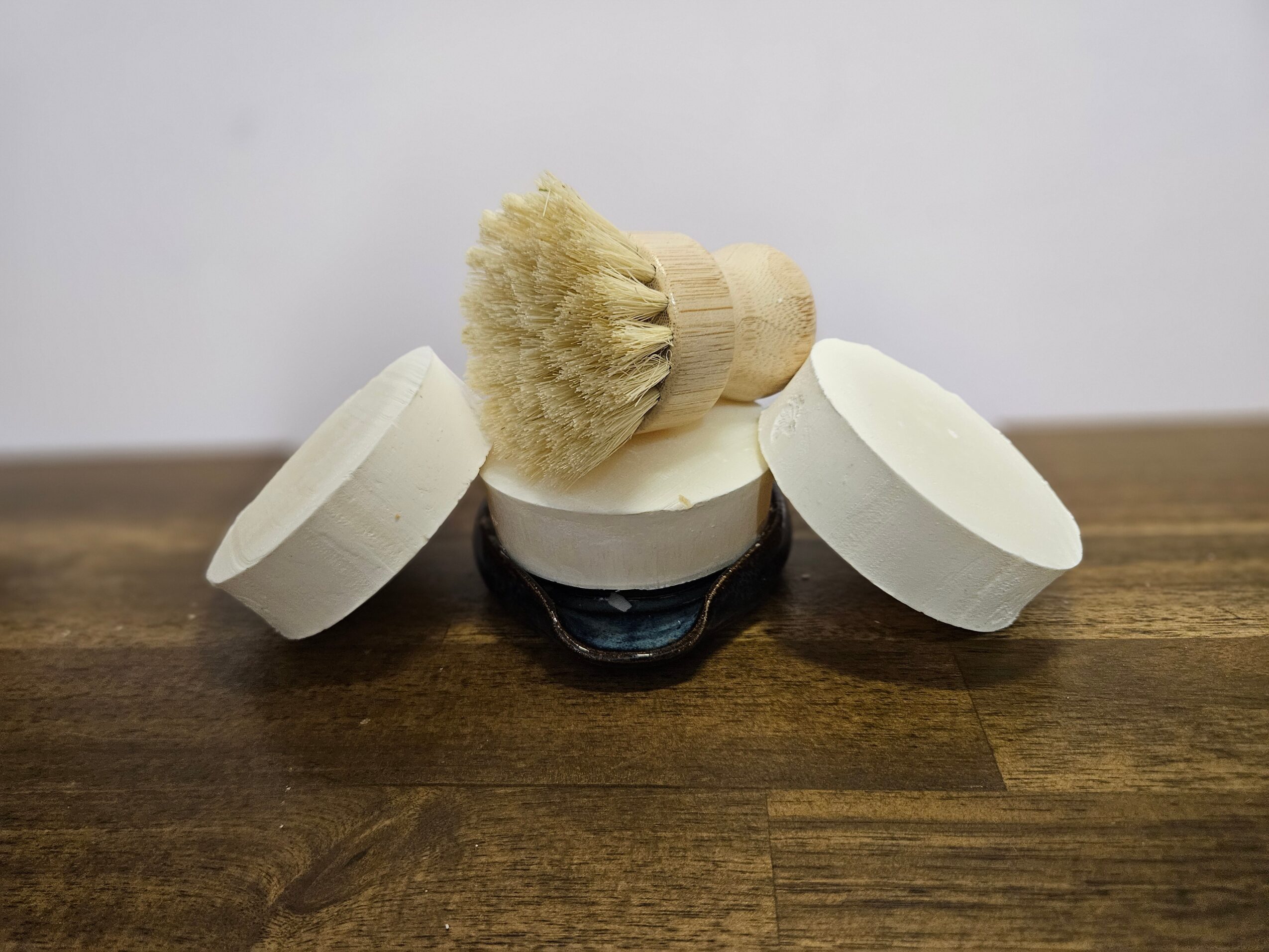 Three white circular soap bars and a wooden brush with a beige bristle head on a wooden surface.