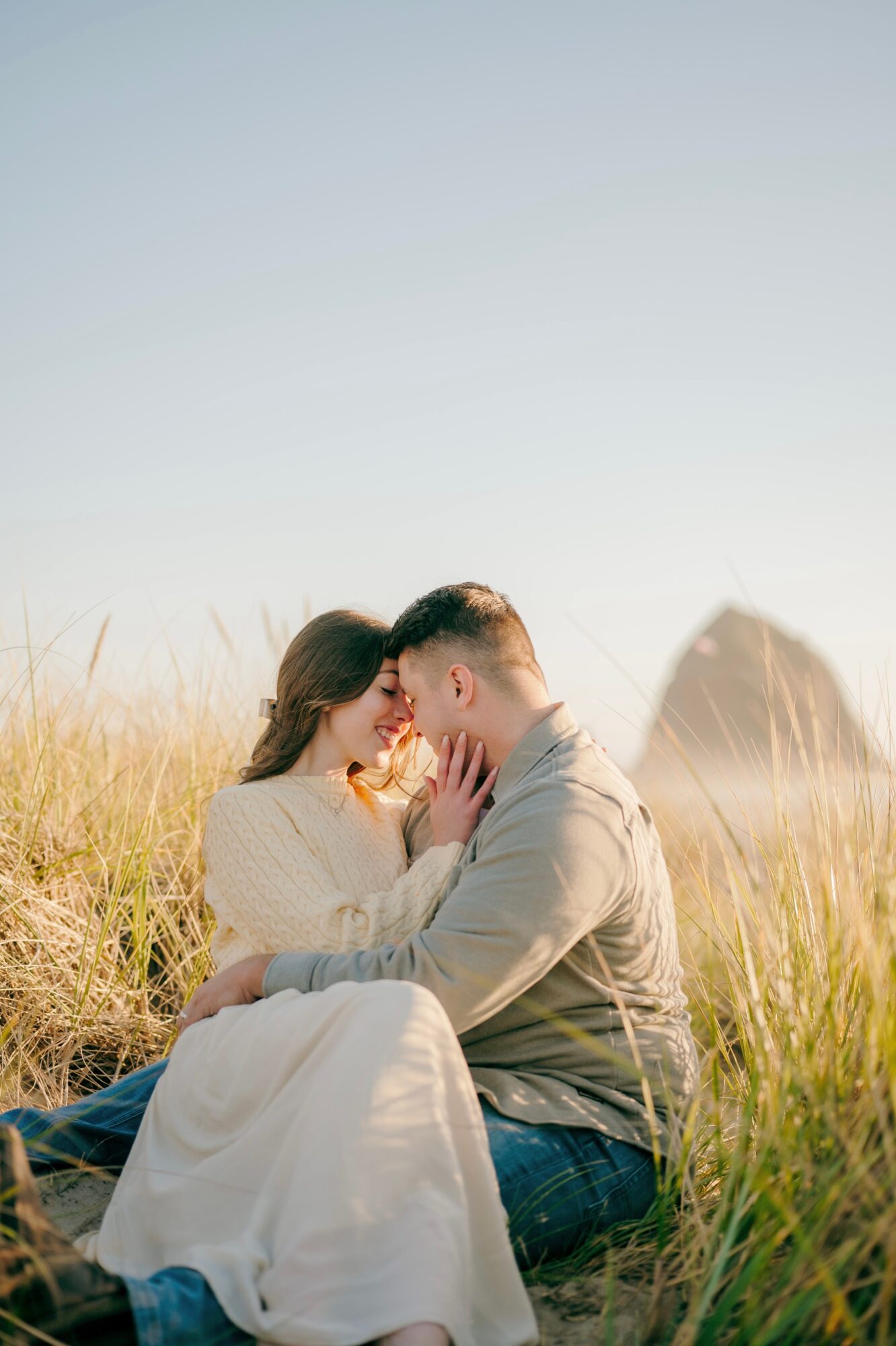 A couple sitting in a grassy field with a large rock in the background, touching foreheads and holding hands.