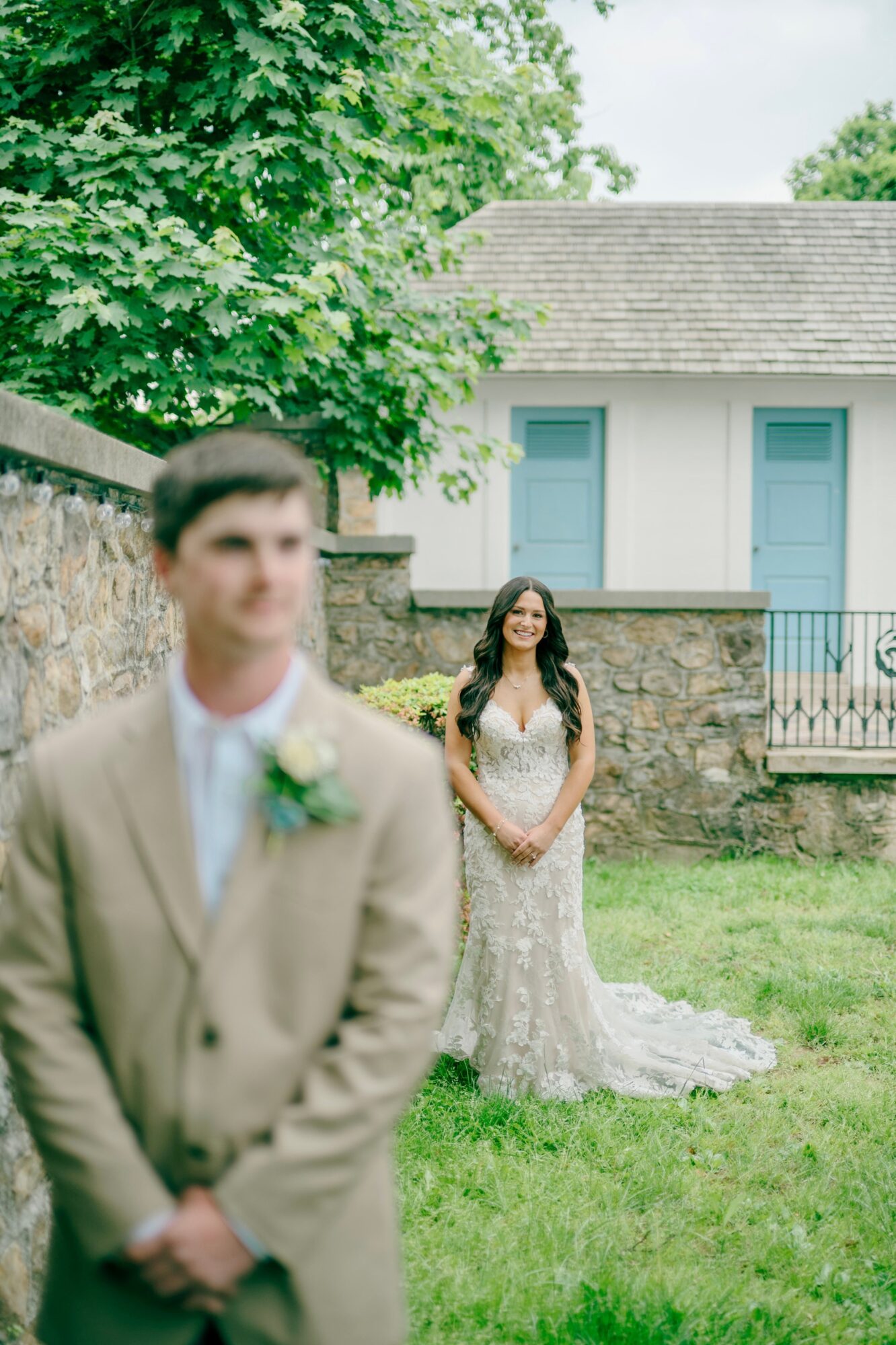 A woman in a wedding dress stands outdoors on grass, smiling, with a stone wall and house in background.