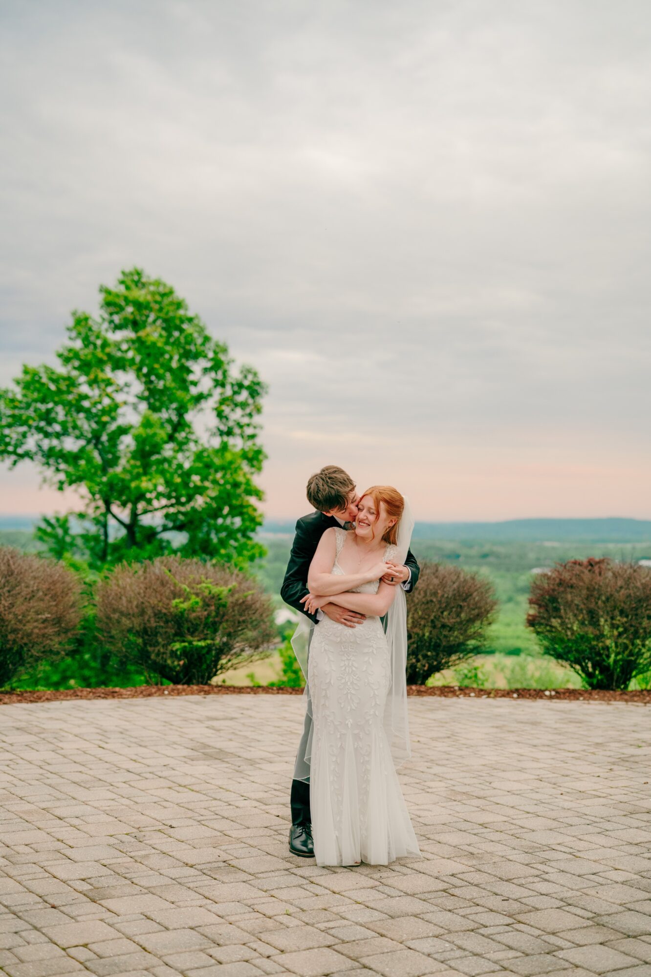 A couple embracing outdoors on a paved area with a tree and landscape in the background.