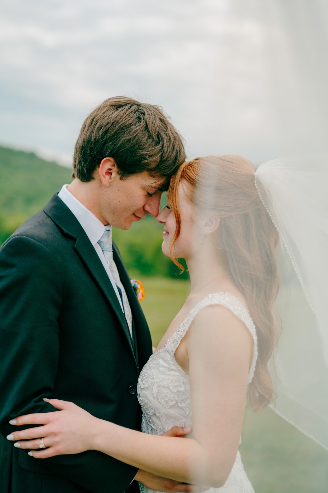 A couple with foreheads touching outdoors, smiling, with a cloudy sky and greenery in the background.
