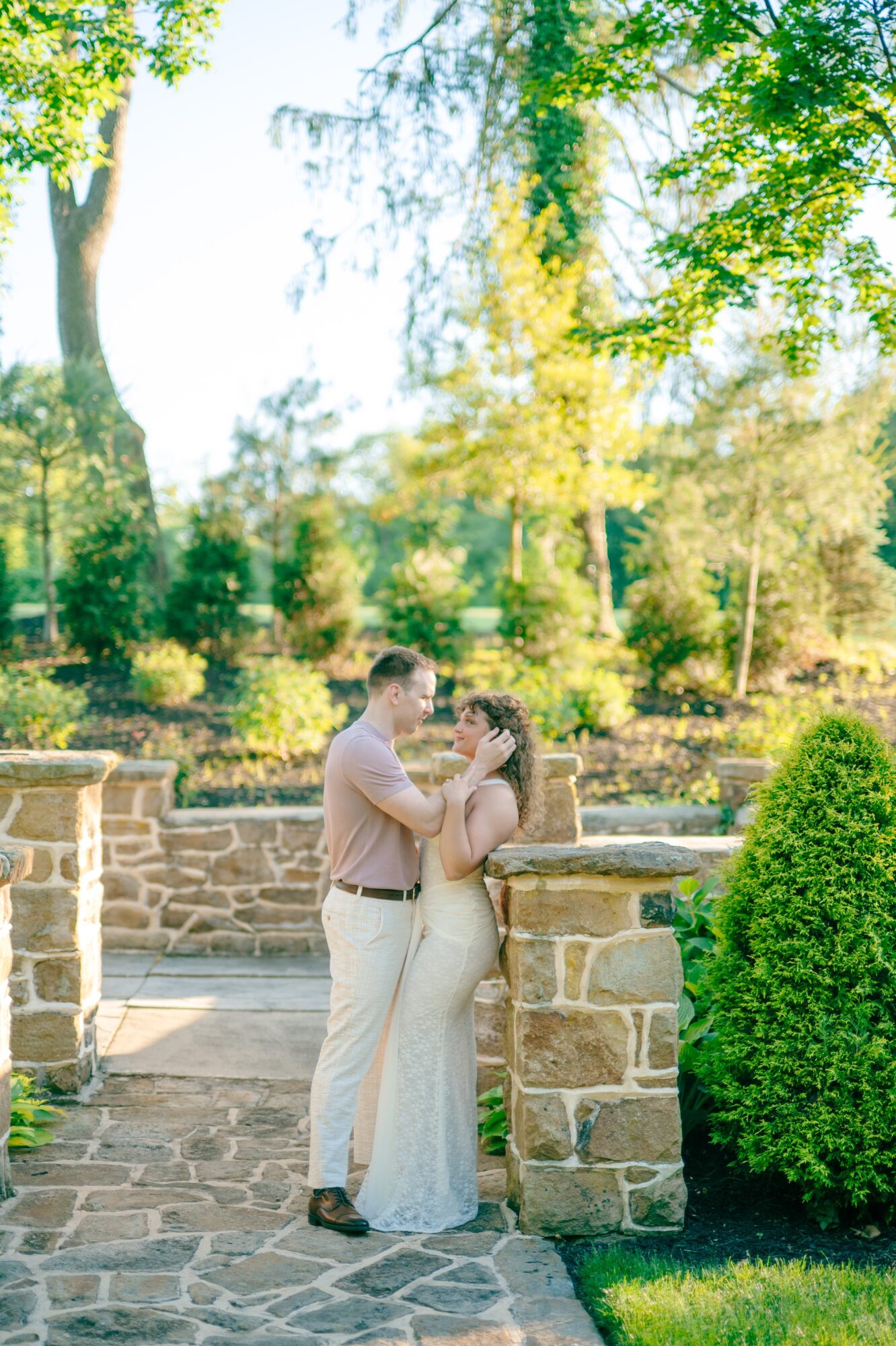 A couple stands close together outdoors, holding hands and looking at each other, surrounded by trees and greenery.