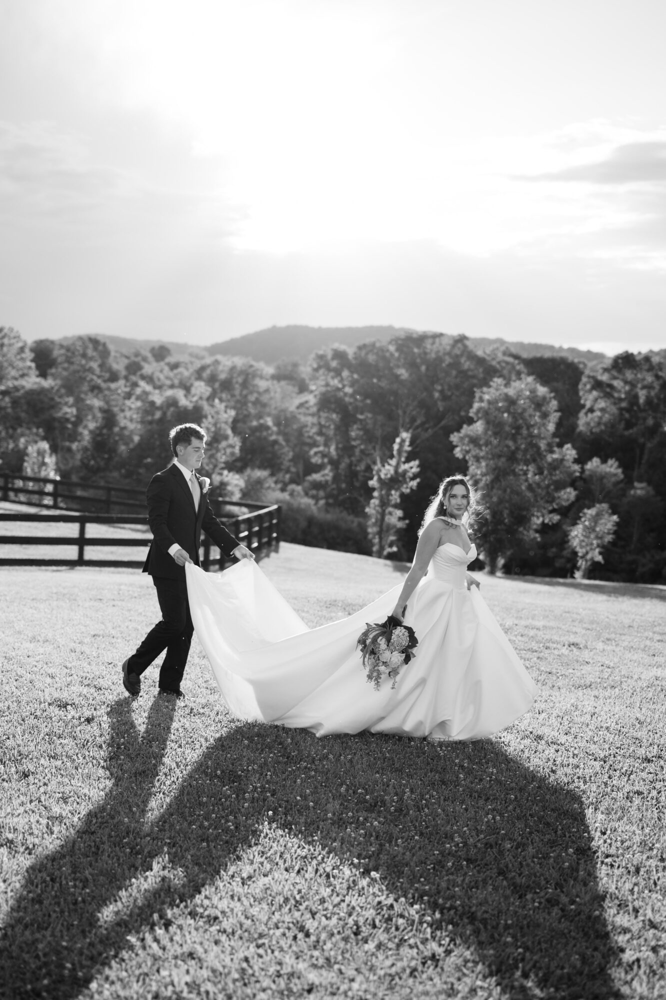 A bride and groom holding a bouquet outdoors with trees and hills in background, sunlight overhead, shadows on grass.