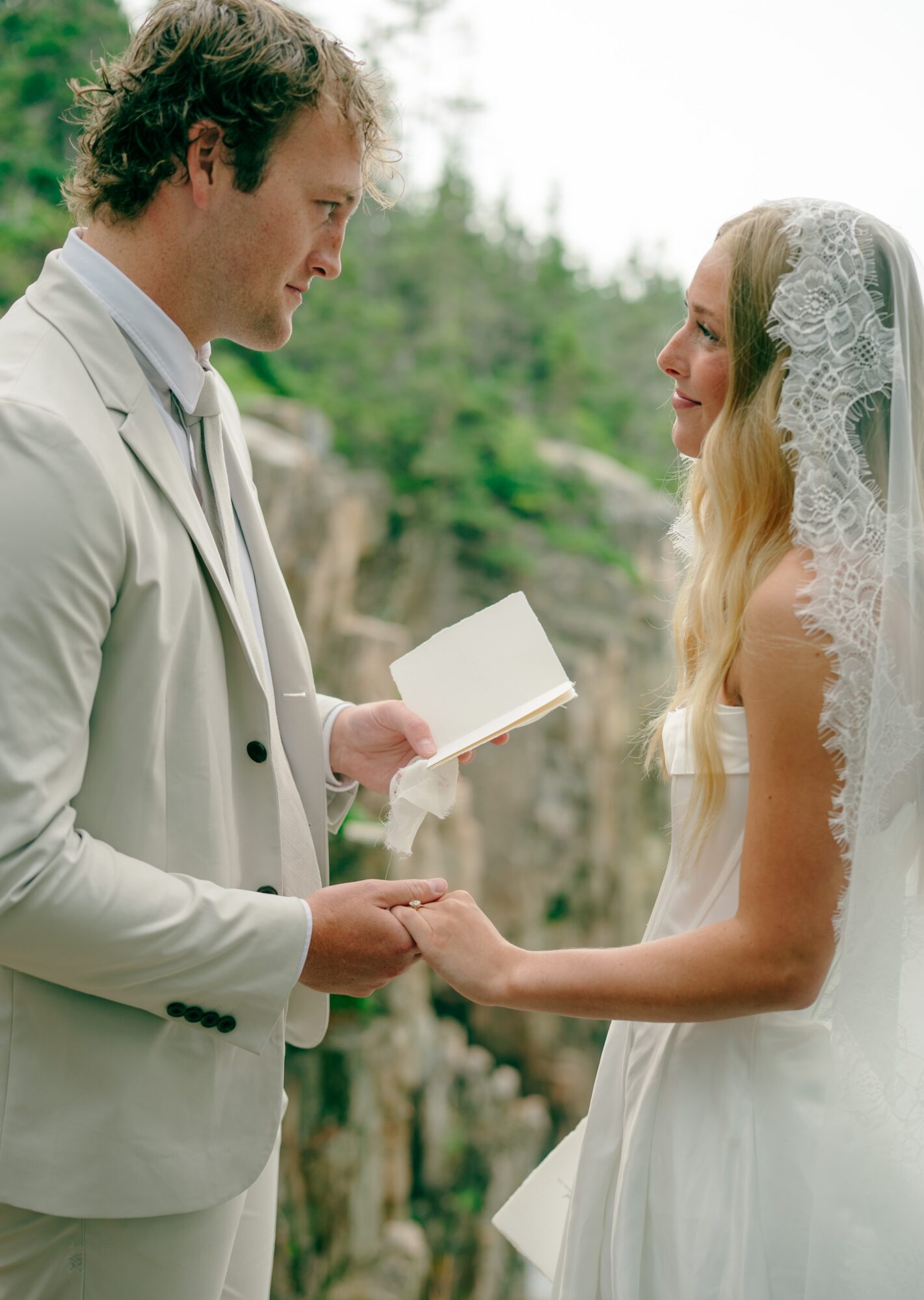 A man and woman holding hands outdoors, with the man reading from a book, both smiling at each other.