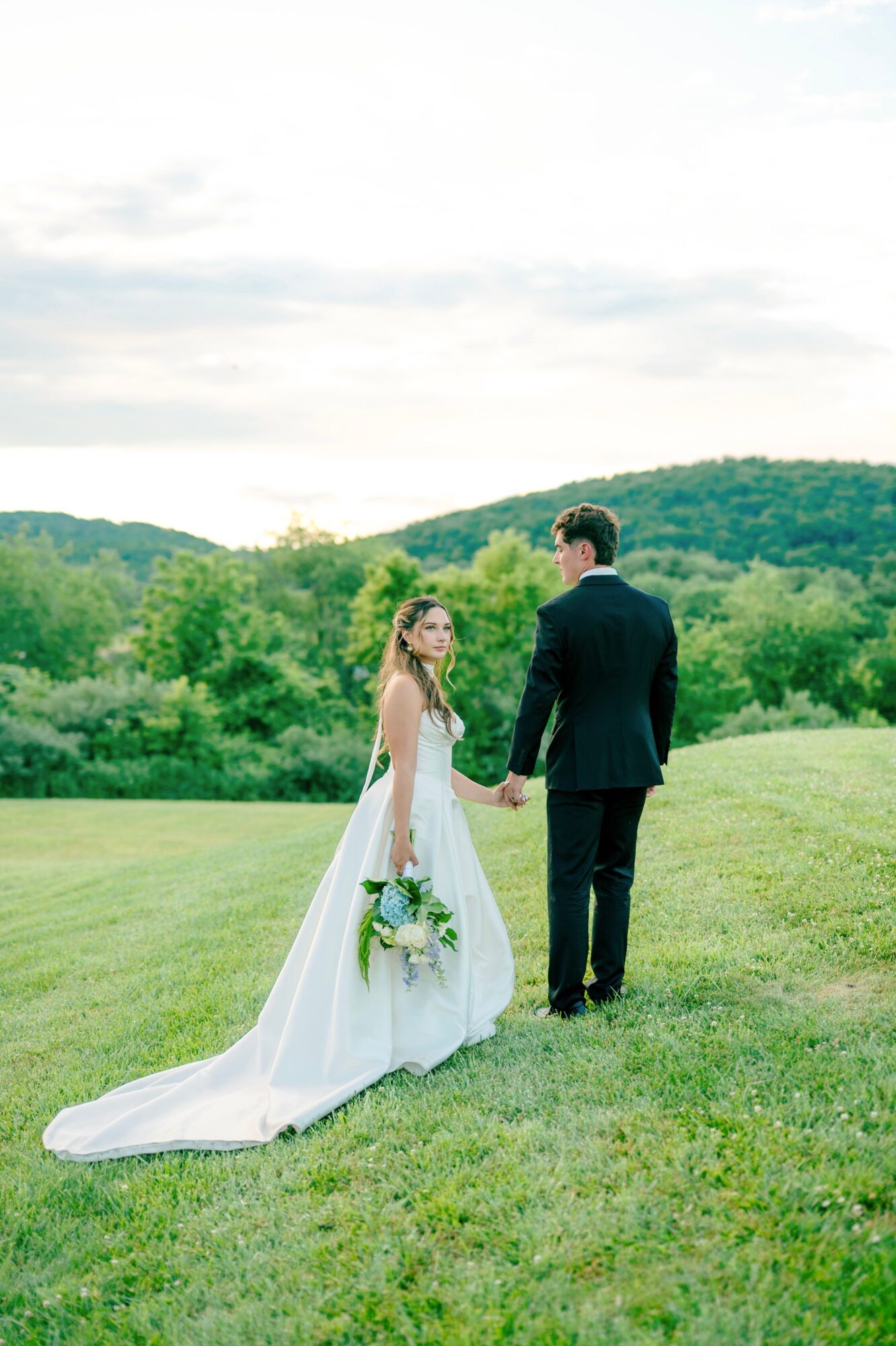 A bride and groom holding hands in a green field with hills and sky in the background.
