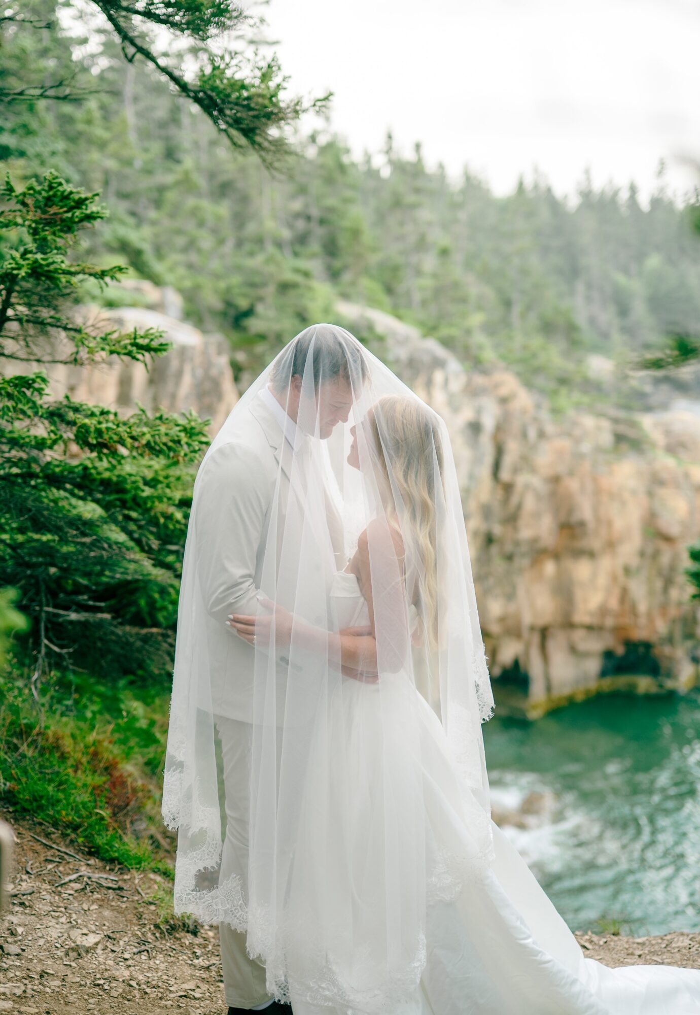 Two people in wedding attire stand close together outdoors near water, with trees and rocks in the background.
