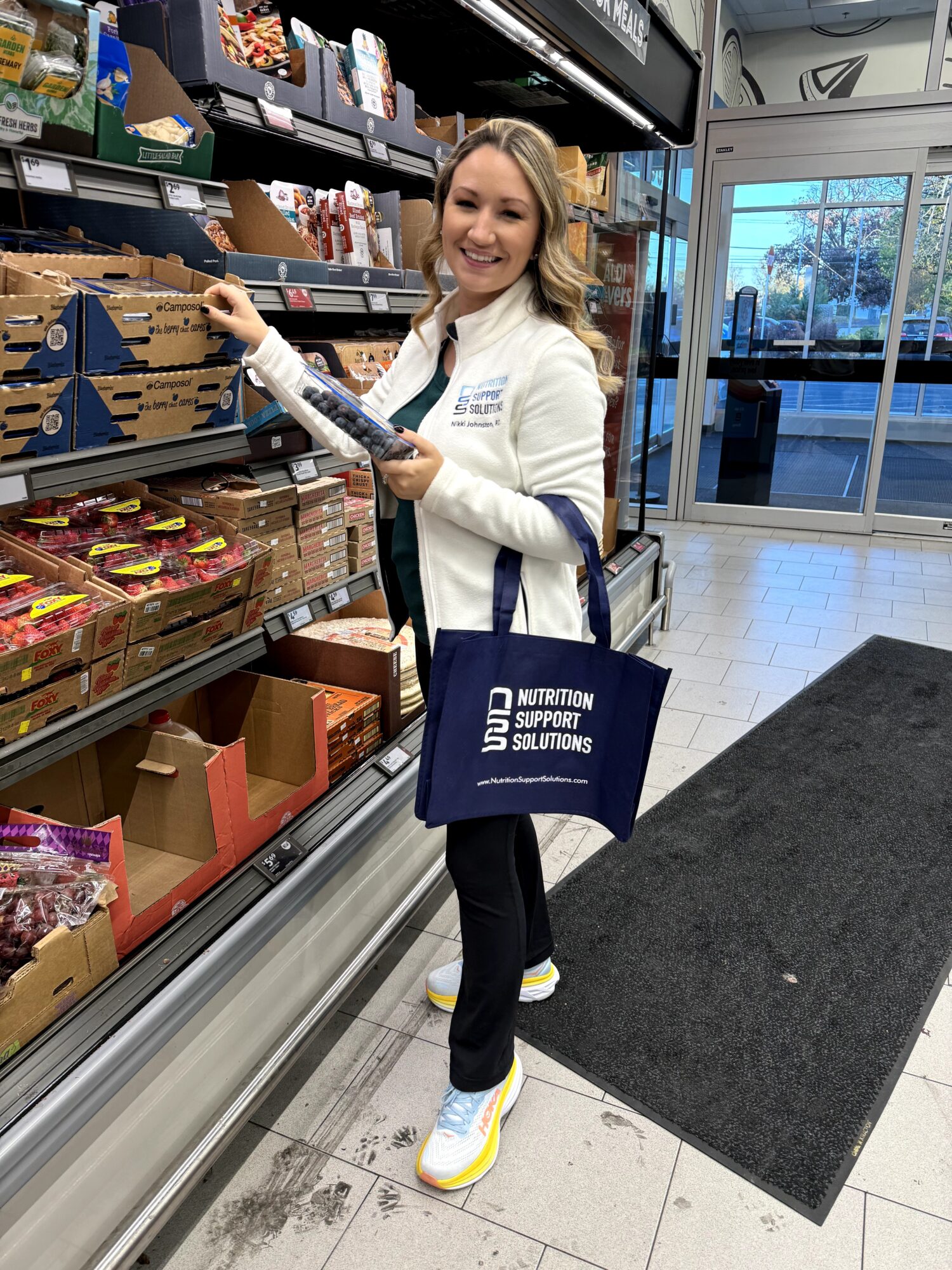 Smiling woman shopping in grocery store aisle, holding a product, with shelves of food items behind her.