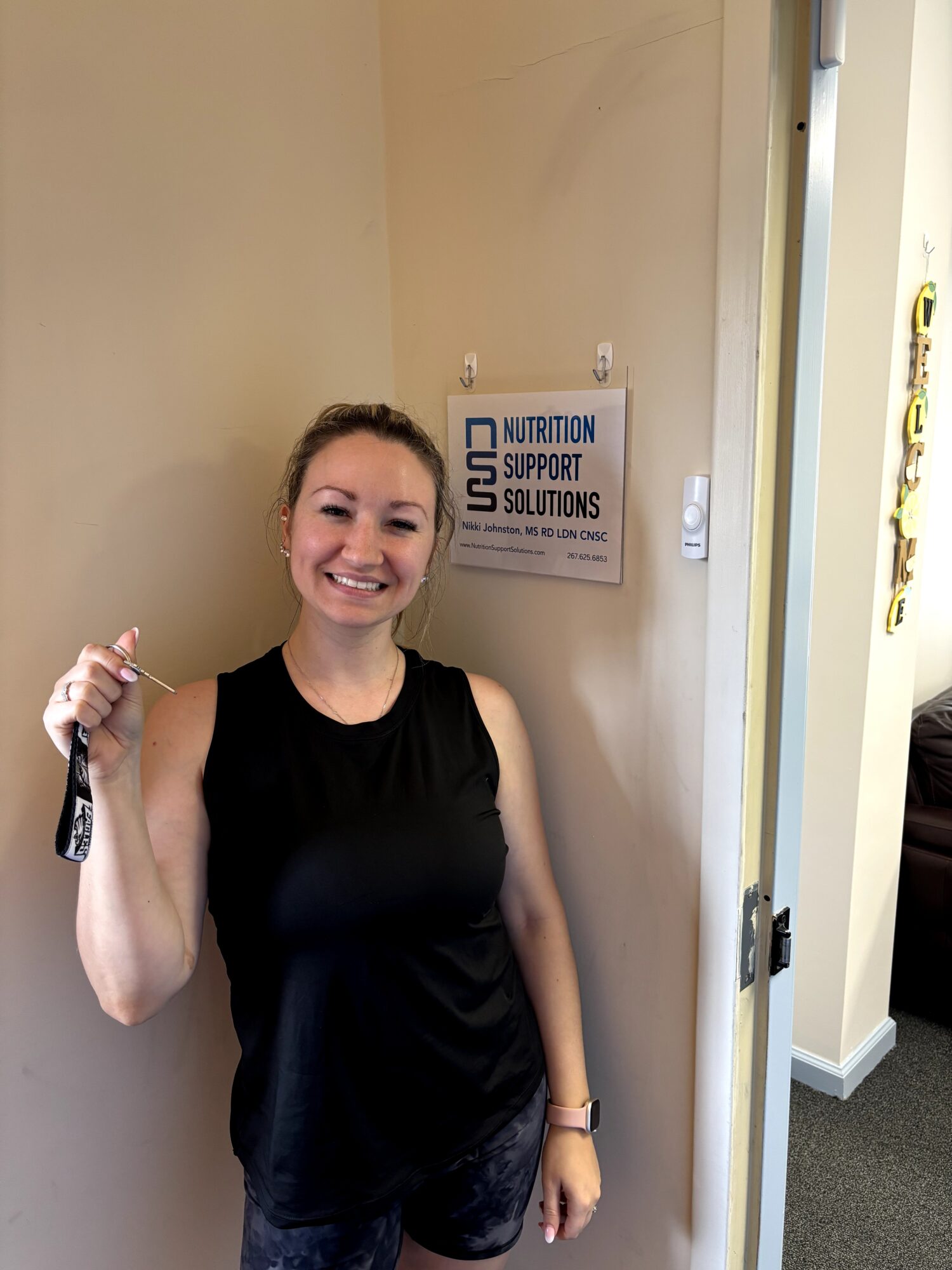 Smiling woman in black sleeveless top holding a key, standing indoors near a sign on the wall.