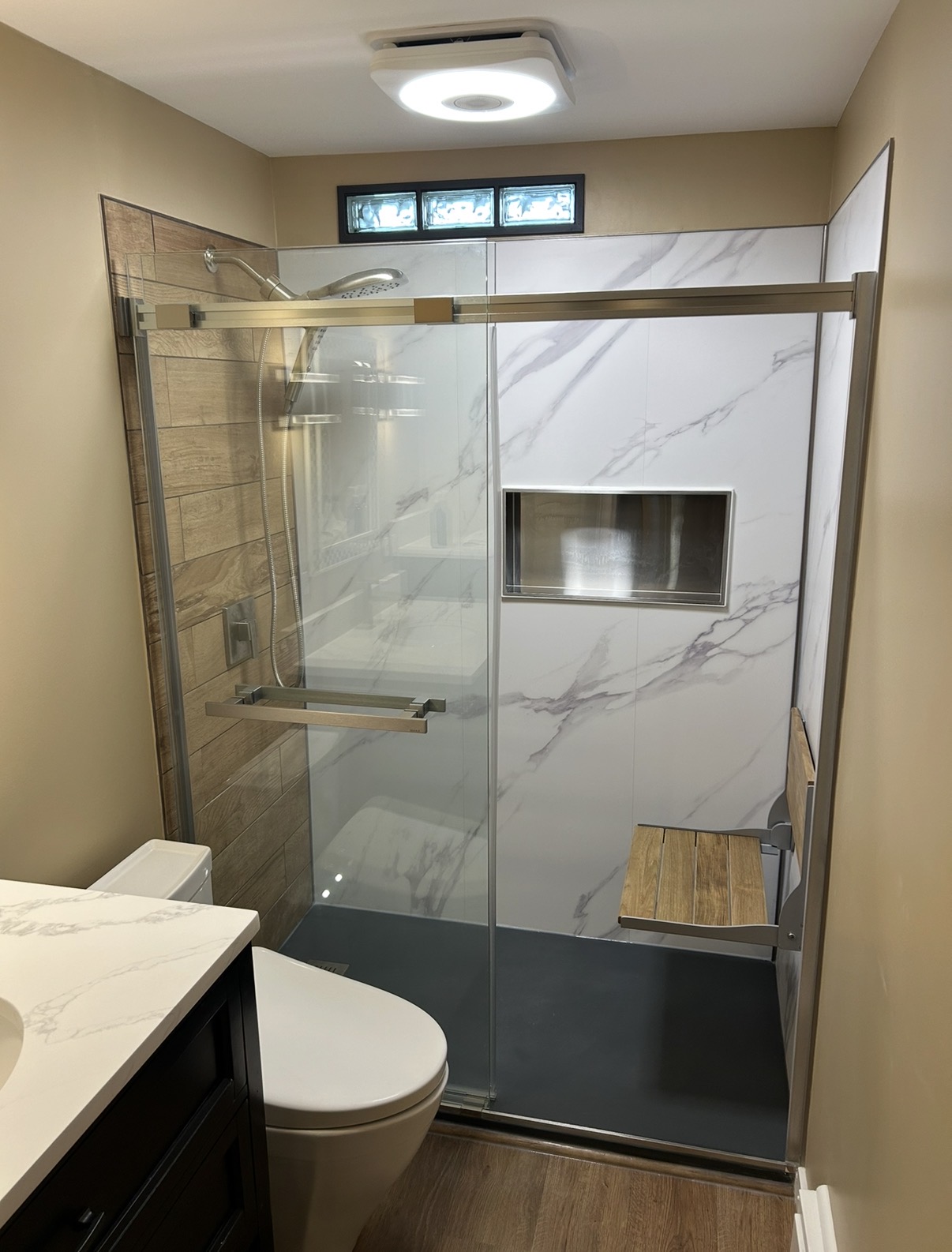 Bathroom shower with glass door, marble wall, small window, and wooden bench inside shower area.