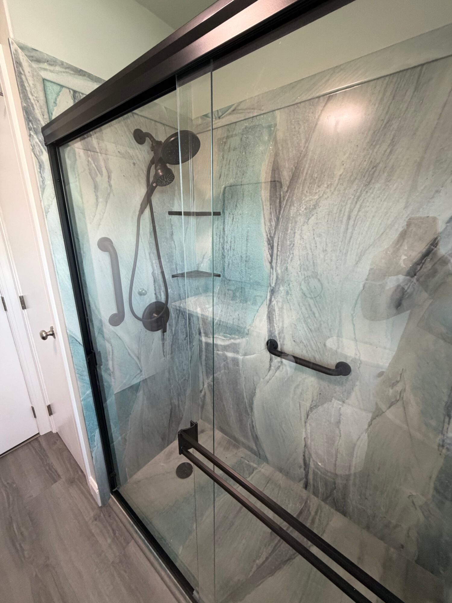 View of a glass shower door with black handles and a black sliding door track, with a marble wall inside the shower.