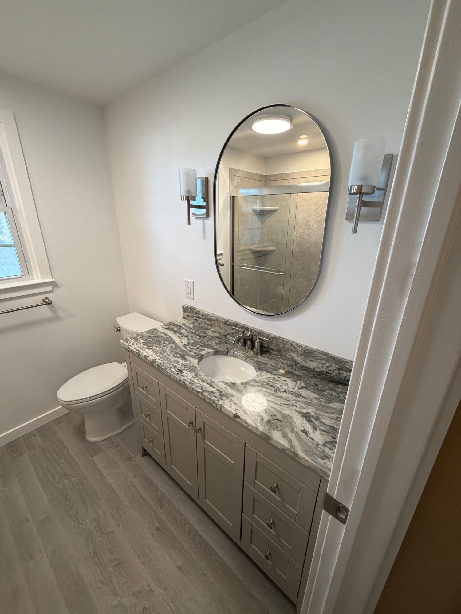 Bathroom with a marble countertop, mirror, and a toilet, viewed from the doorway, with a window and light-colored flooring.