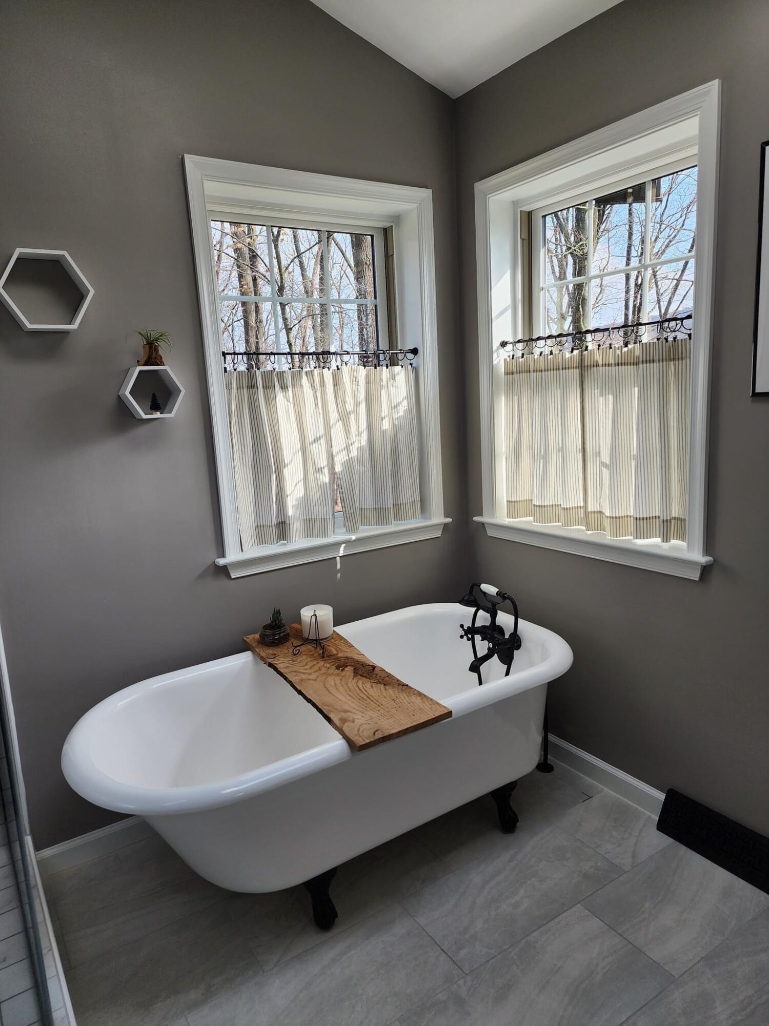 Bathroom with a clawfoot bathtub near two windows, gray walls, and small decorative shelves, natural light coming in.