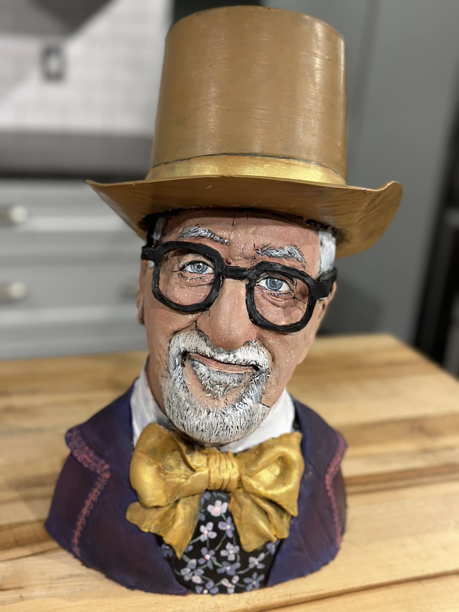 A figurine of a man with glasses, a beard, and a mustache, wearing a large top hat, a purple coat, and a gold bow tie, on a wooden surface.