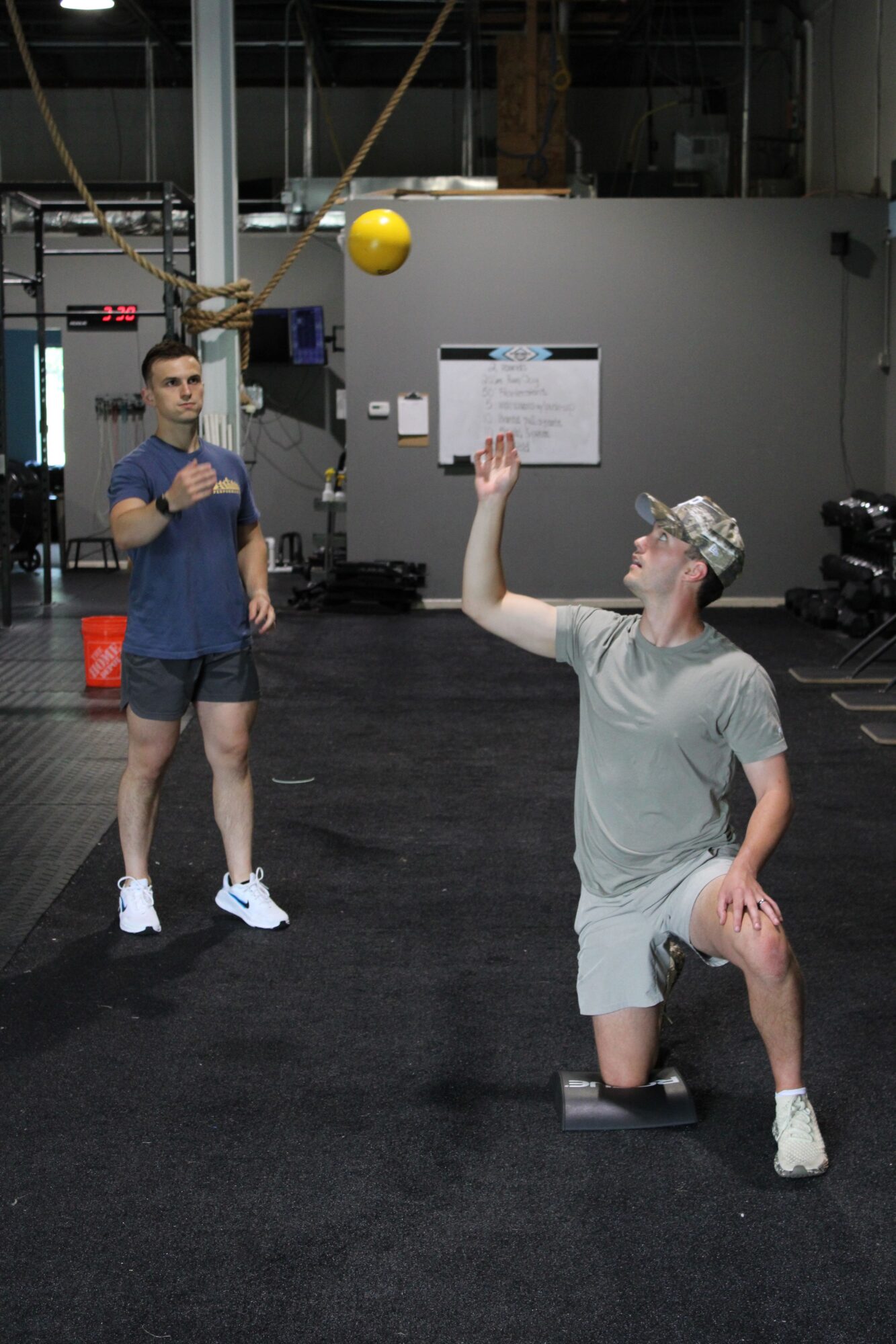 Two women standing in a gym, one holding a whiteboard, the other with hands on hips, black carpeted floor, gym equipment in background.