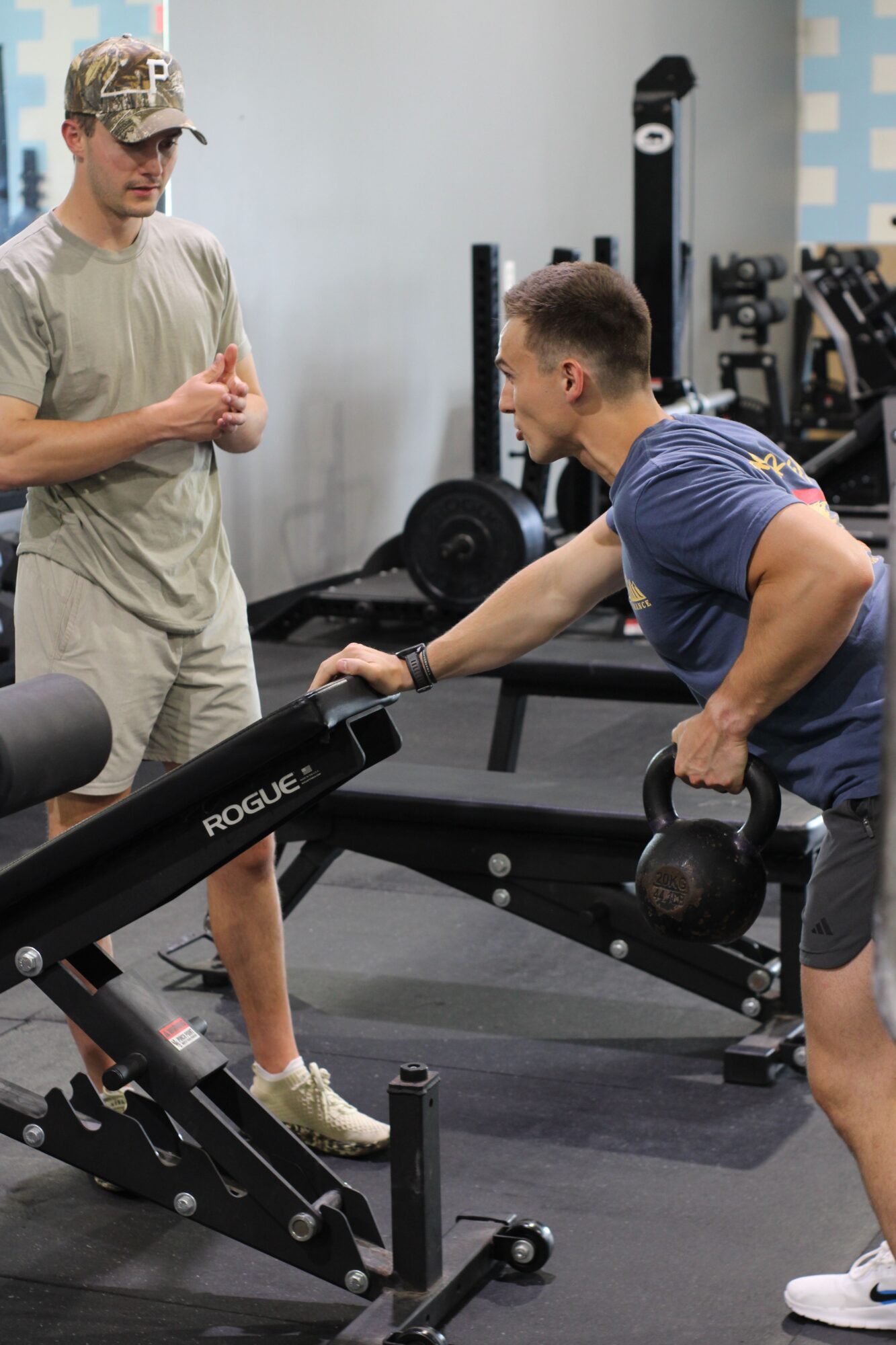 Person lifting a dumbbell while another person stands nearby in a gym setting.