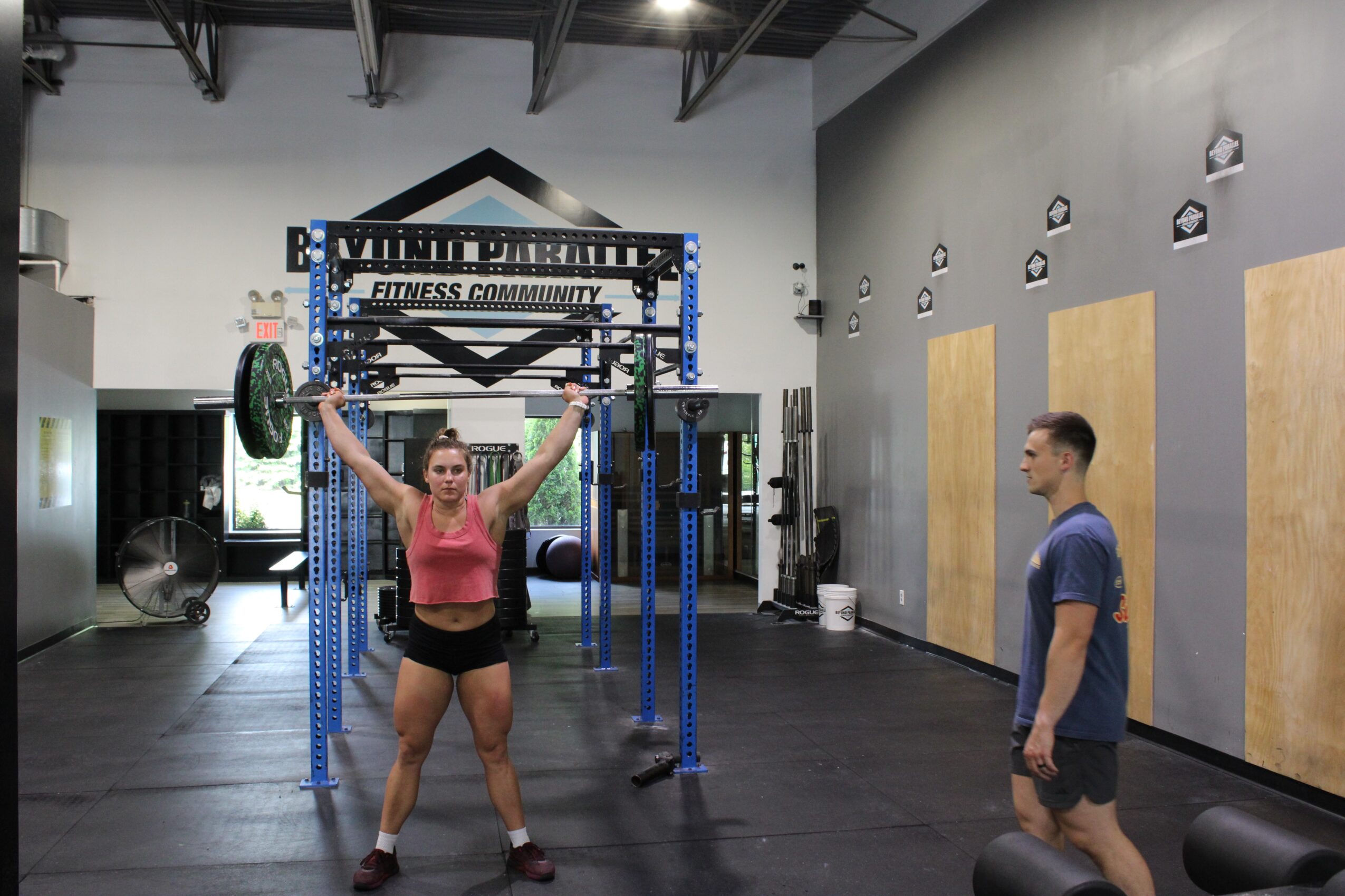 Woman lifting a barbell overhead in a gym with a man watching nearby, gym equipment in background.
