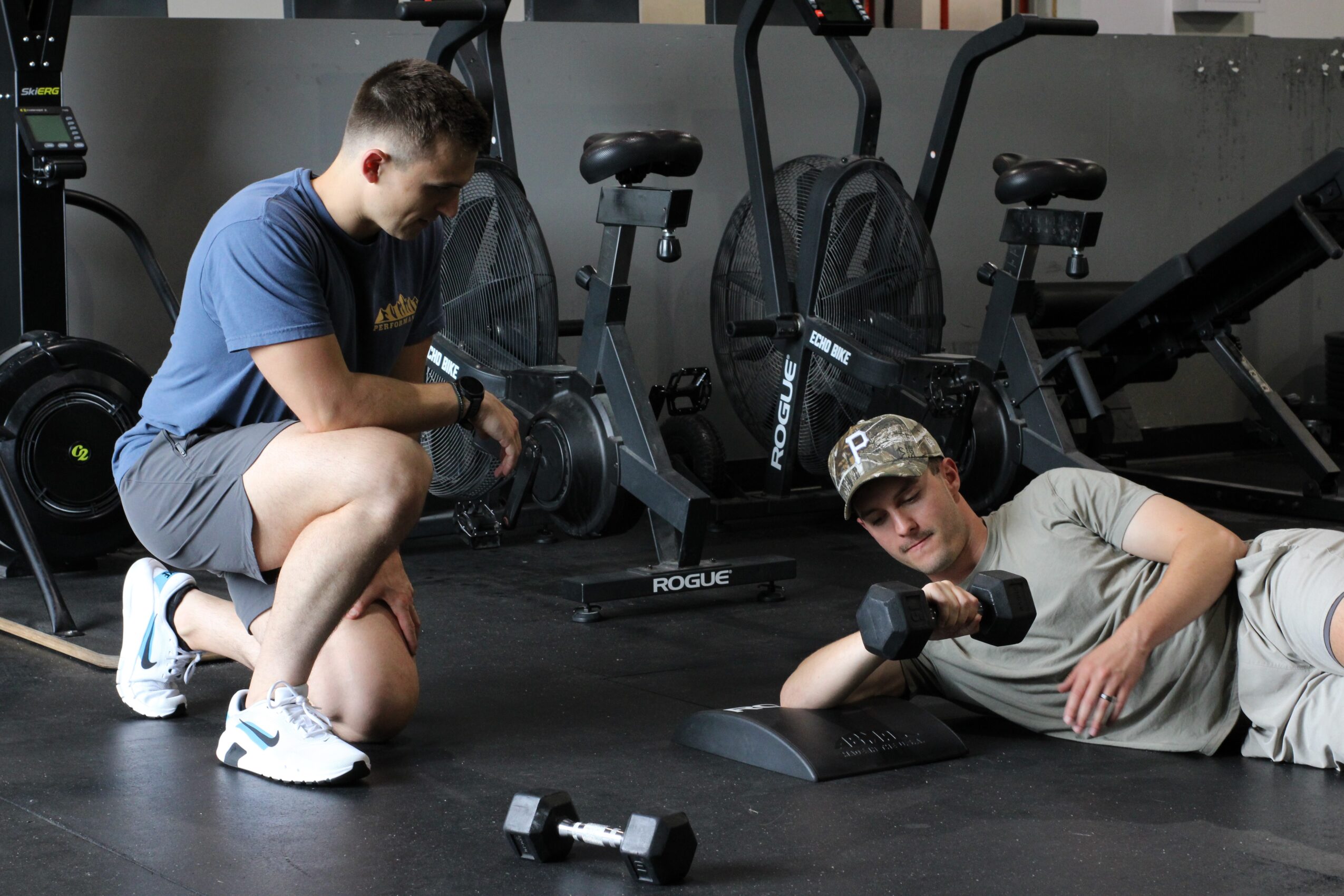 Two young men exercising with dumbbells in a gym, one kneeling and the other lying on the floor, surrounded by gym equipment.