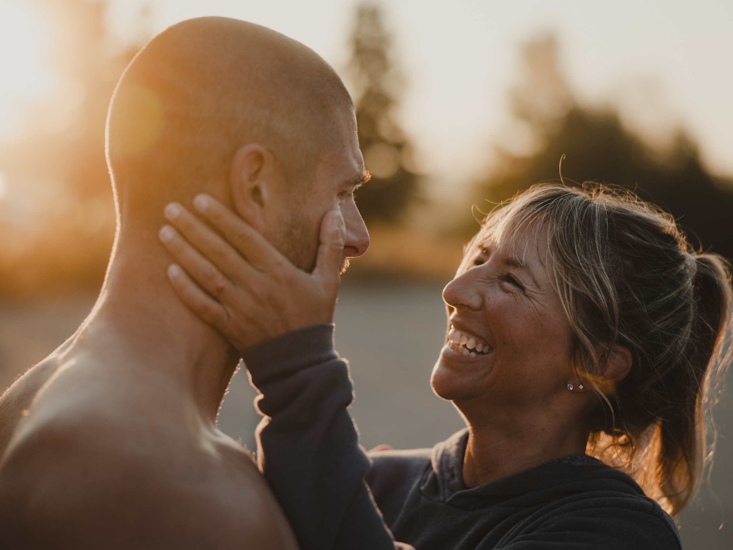 Woman smiling and holding man's face outdoors during sunset, both looking at each other.