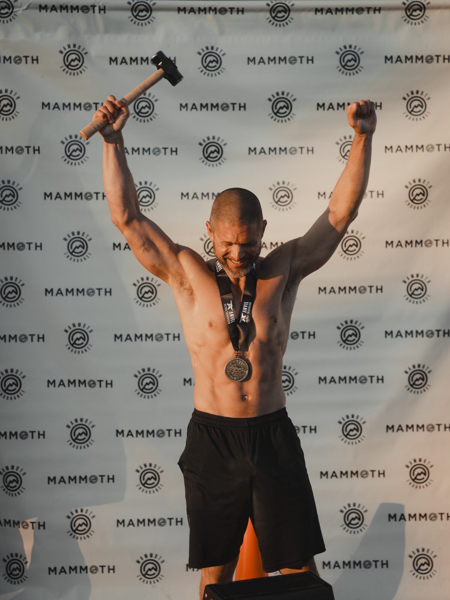 Male athlete with a medal around his neck, holding a trophy in each hand, arms raised in victory, shirtless, in front of a patterned backdrop.