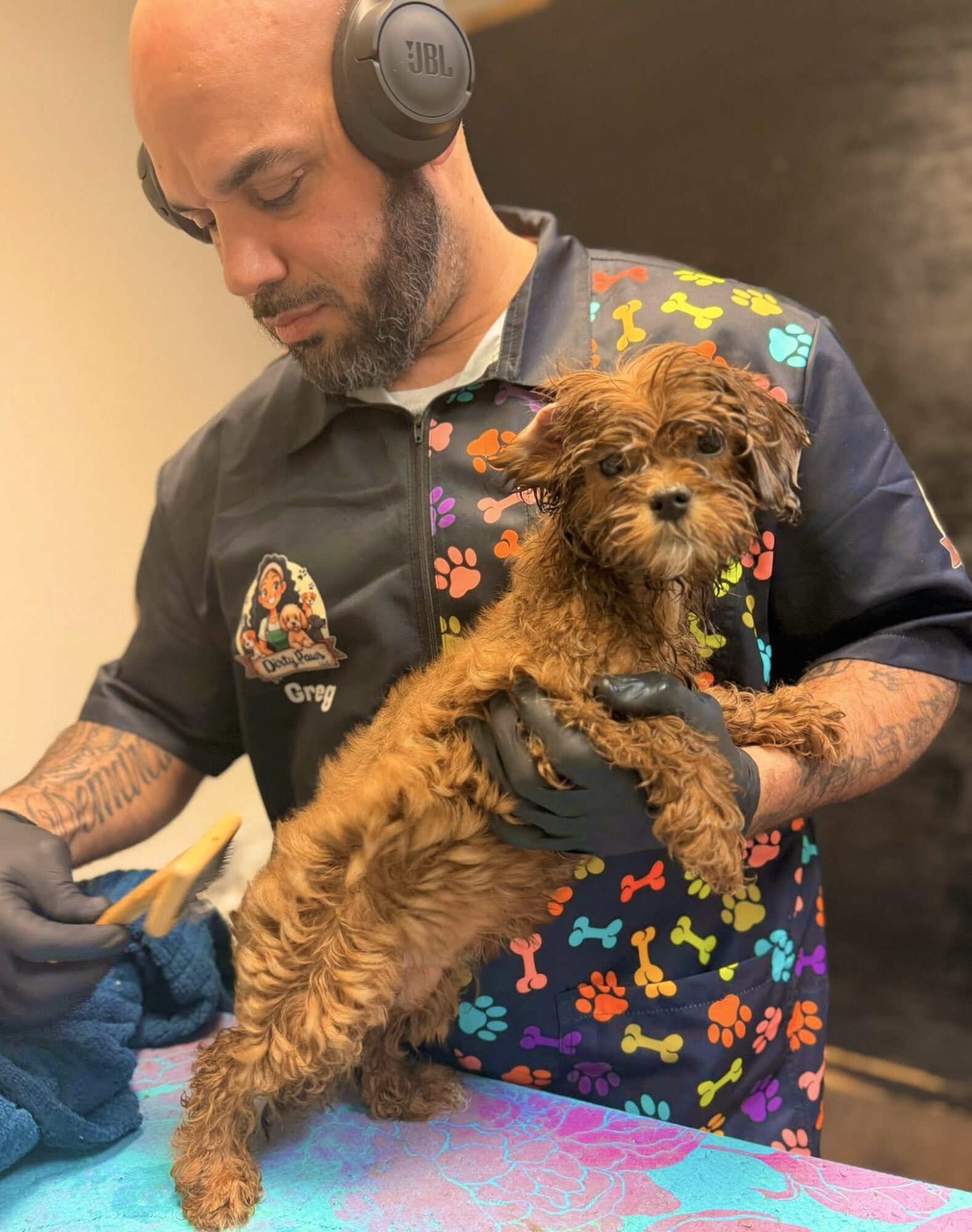 Man with headphones holding a small, curly-haired puppy, wearing a colorful shirt with bone and paw print patterns.