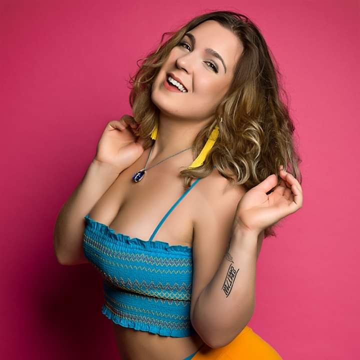 Smiling woman with curly hair, wearing a blue top and yellow earrings, against a pink background.