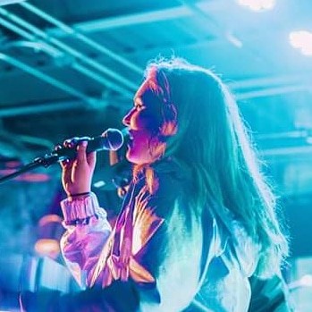 Young woman singing into a microphone on stage with colorful lighting, long hair, and a jacket, facing left.