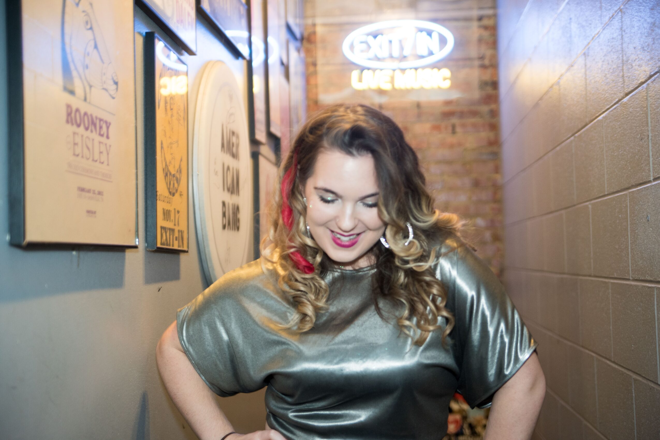 Woman with wavy hair and red earrings smiling, standing in a hallway with posters on the wall and a neon sign in the background.