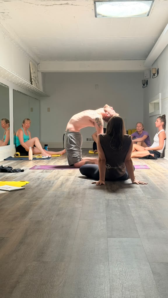 Group of people practicing yoga in a bright room with a wooden floor, some sitting and some stretching, with a person in the center assisting.