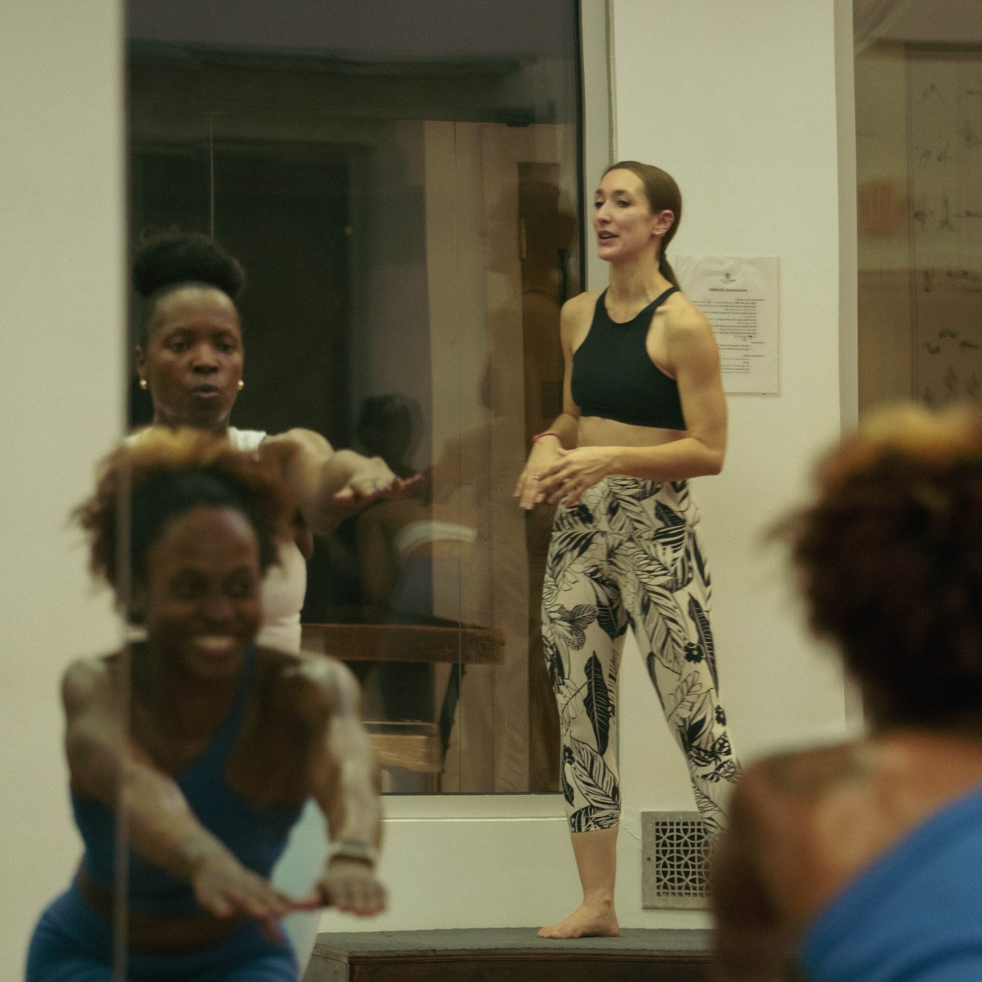 Group of women in a room, one standing and speaking, others sitting and smiling, with a window and wall behind.