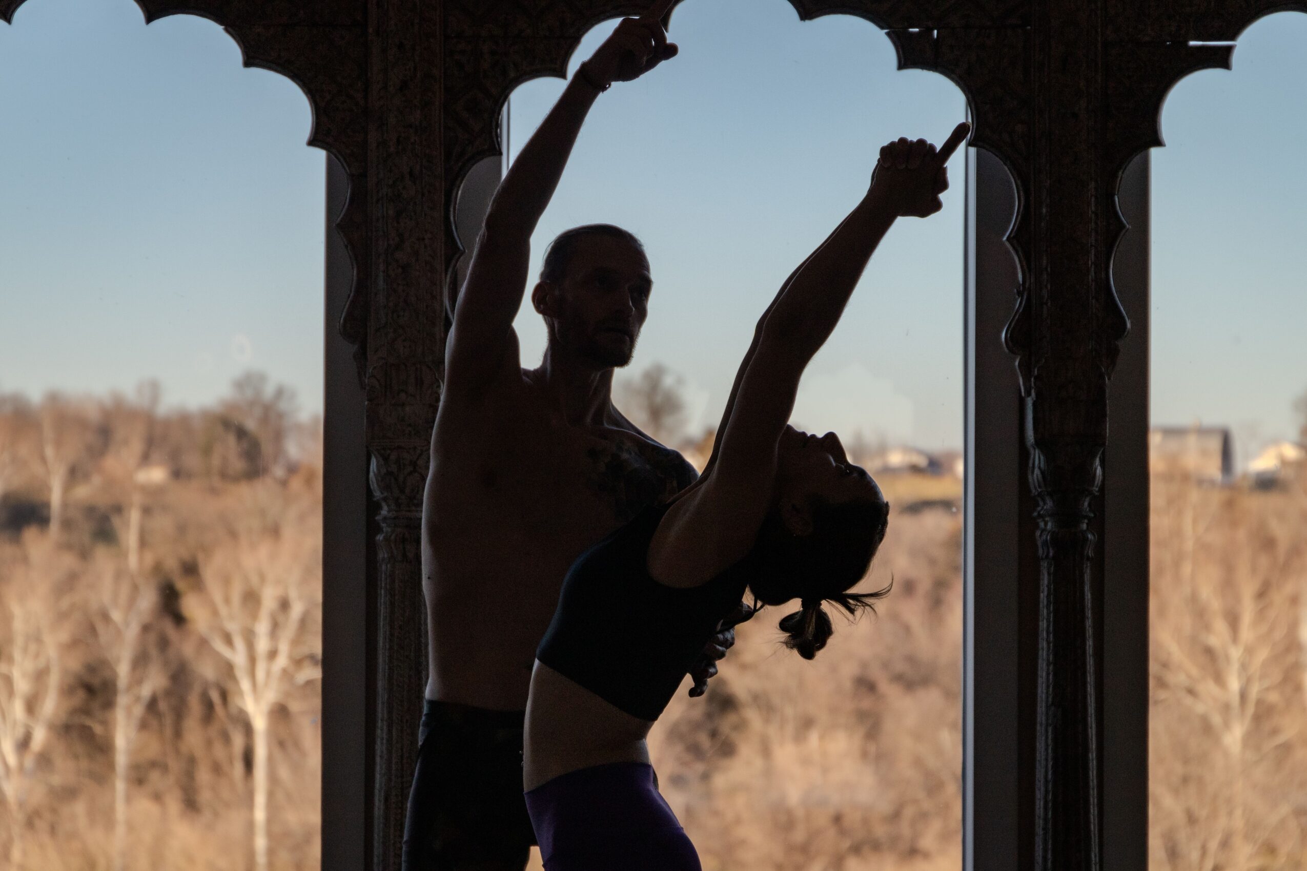 Two people perform a dance pose under an ornate archway with a landscape background.