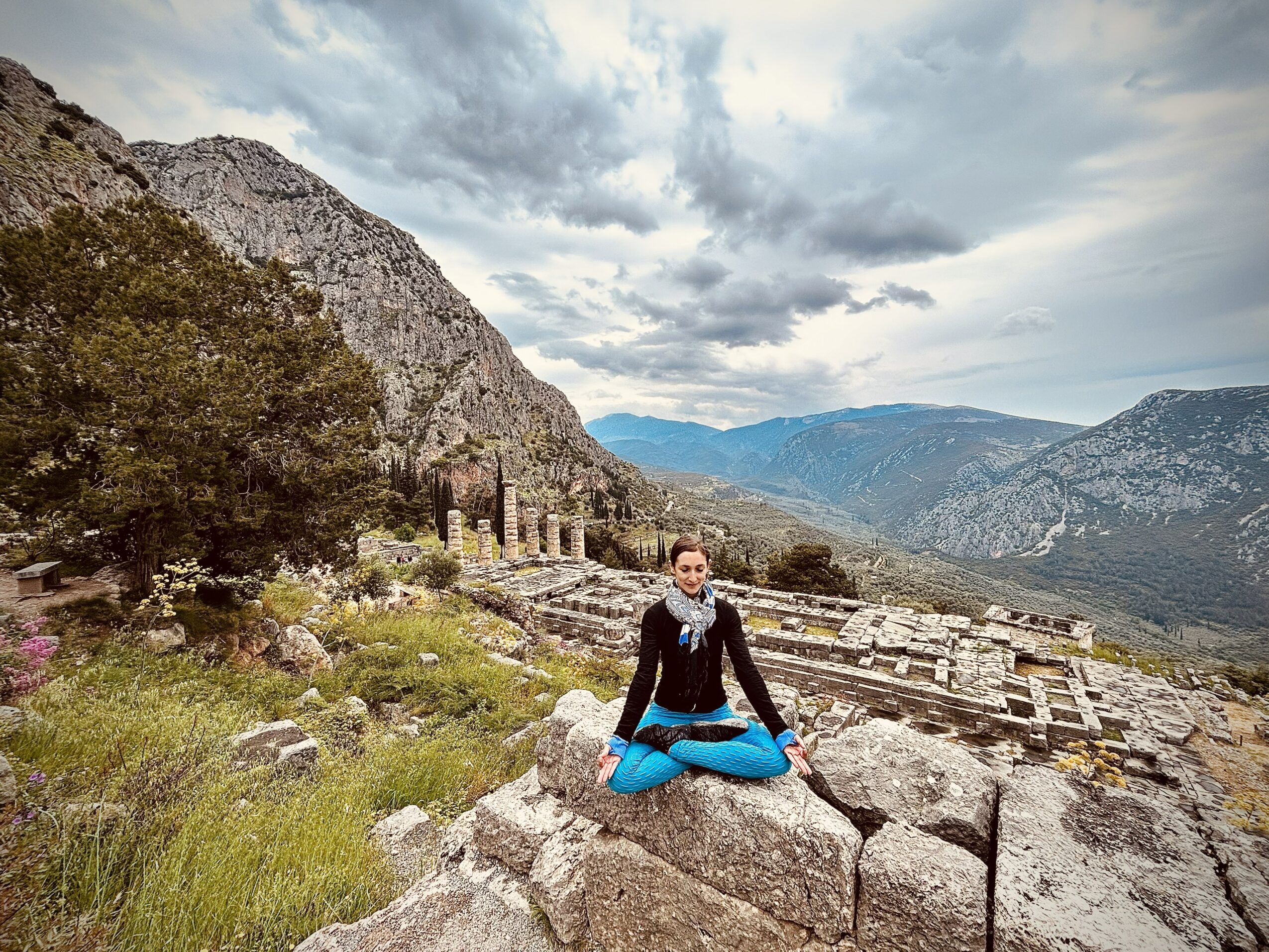 Person sitting cross-legged on a large rock in a mountainous landscape with ruins and cloudy sky.