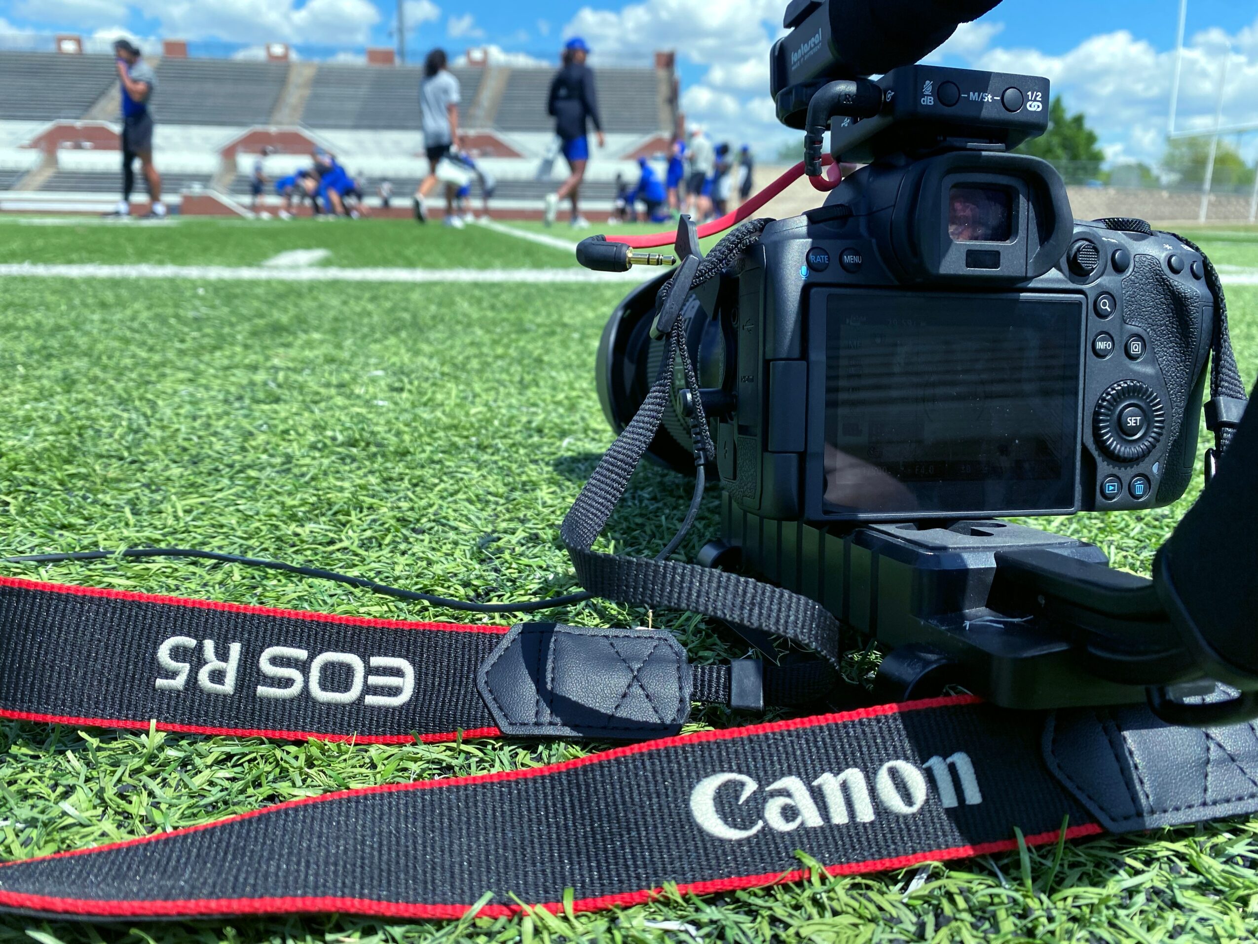 Camera on grass field with people in the background, blue sky, and clouds, capturing a sports event.