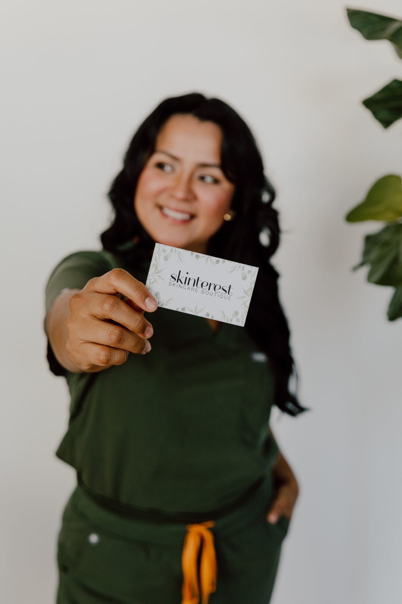 Woman with dark hair smiling, holding a card, standing near a plant, wearing a green outfit, against a plain background.