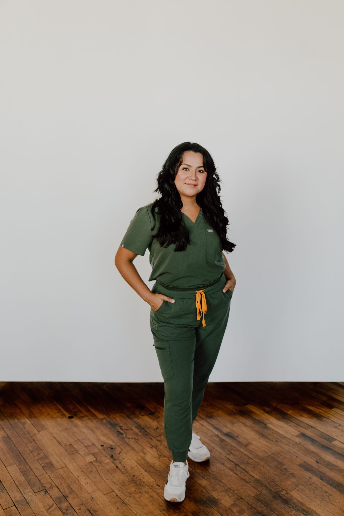 Woman with long dark hair in green scrubs standing with hands in pockets against a plain white wall.