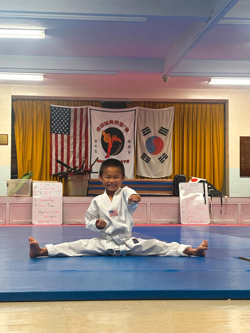 Child in martial arts uniform performing a split on blue mat, smiling, with flags and curtains in background.