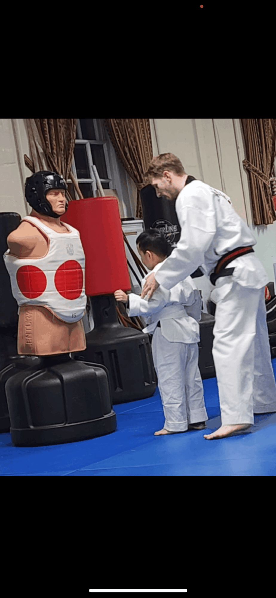 Martial arts instructor teaching children with protective gear in a training room, blue mat, curtains, and window in background.