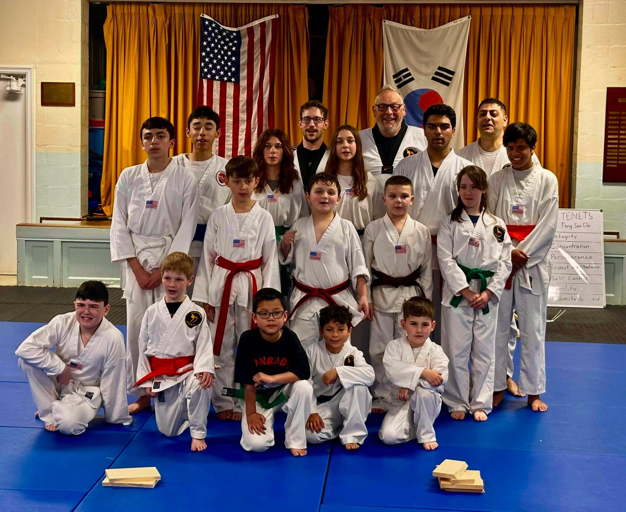 Group of children and adults in martial arts uniforms posing on a blue mat, with American and South Korean flags behind them.