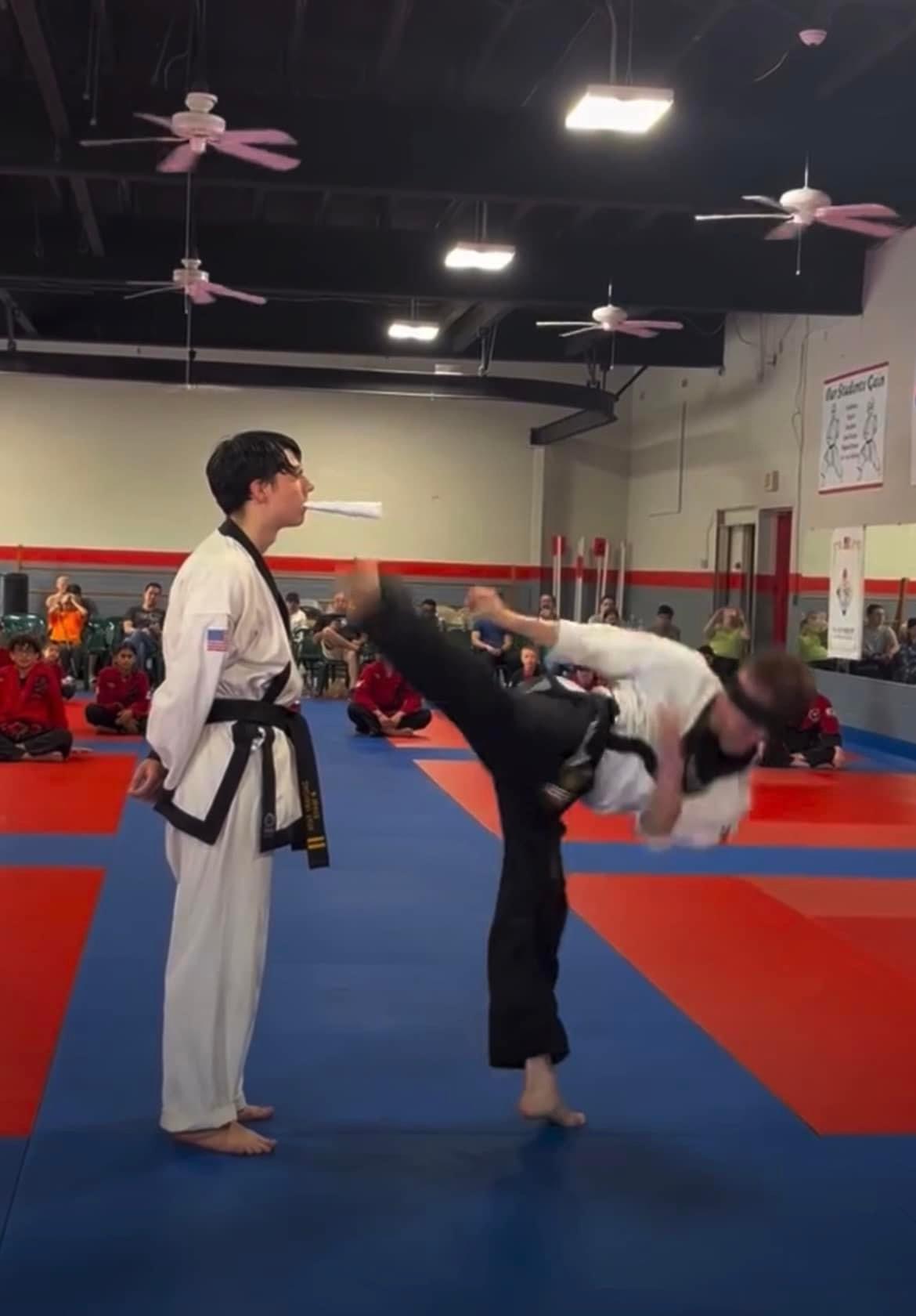 Two martial artists practicing in a dojo, one performing a high kick on the other, with people and equipment in background.