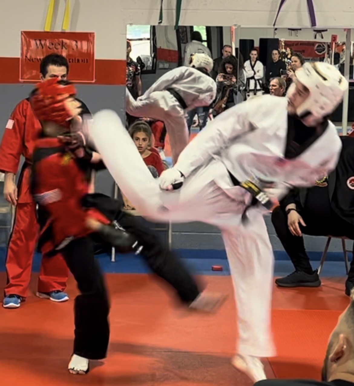 Two martial artists sparring, one in red and the other in white, with spectators watching in the background.