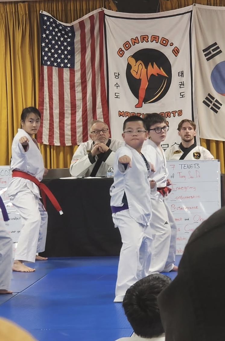 Children in martial arts uniforms perform in front of judges, with flags and banners in the background, on a blue mat.